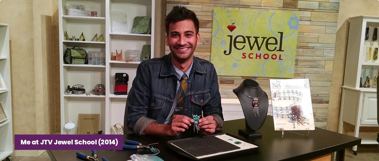 A man sitting at a jewelry designing table at JTV Jewel School, holding a turquoise-colored jewelry piece, surrounded by jewelry making tools and displays of jewelry. The background features shelves with jewelry supplies and a sign that reads 'jewel SCHOOL'.