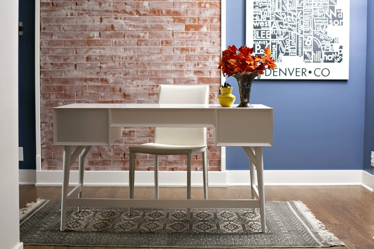 Interior of a room featuring a white desk with a matching chair, a vase of red flowers, a yellow decorative item, a brick accent wall, and a framed art piece on a blue wall.