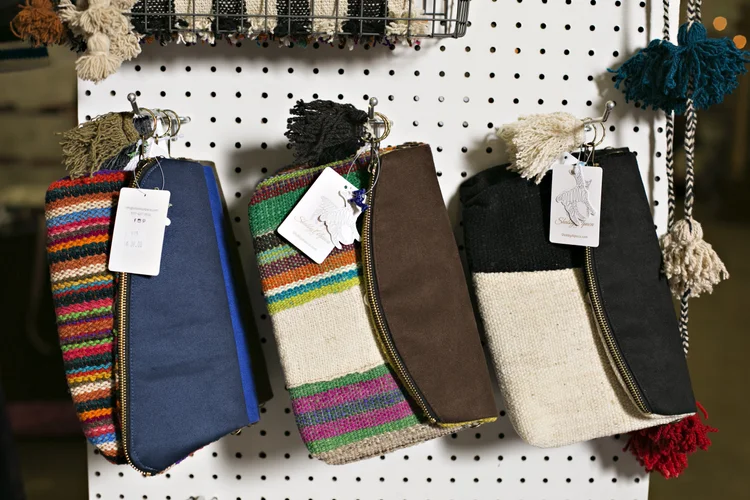 Display of four colorful hand-knit bags with zippers and pom-pom tassels, hanging on a pegboard.