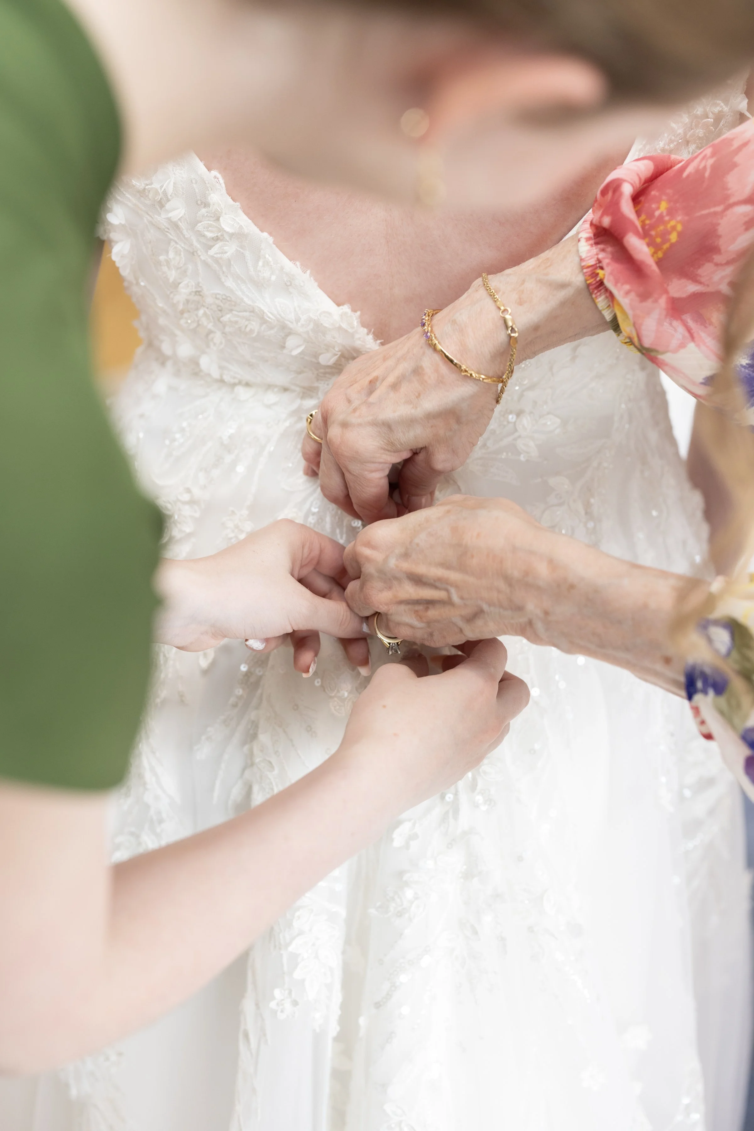 An elderly woman dressed in a white lace wedding gown is being assisted with her dress. Multiple hands are helping her, and she is wearing a gold bracelet and ring. The scene appears to be part of a wedding preparation.