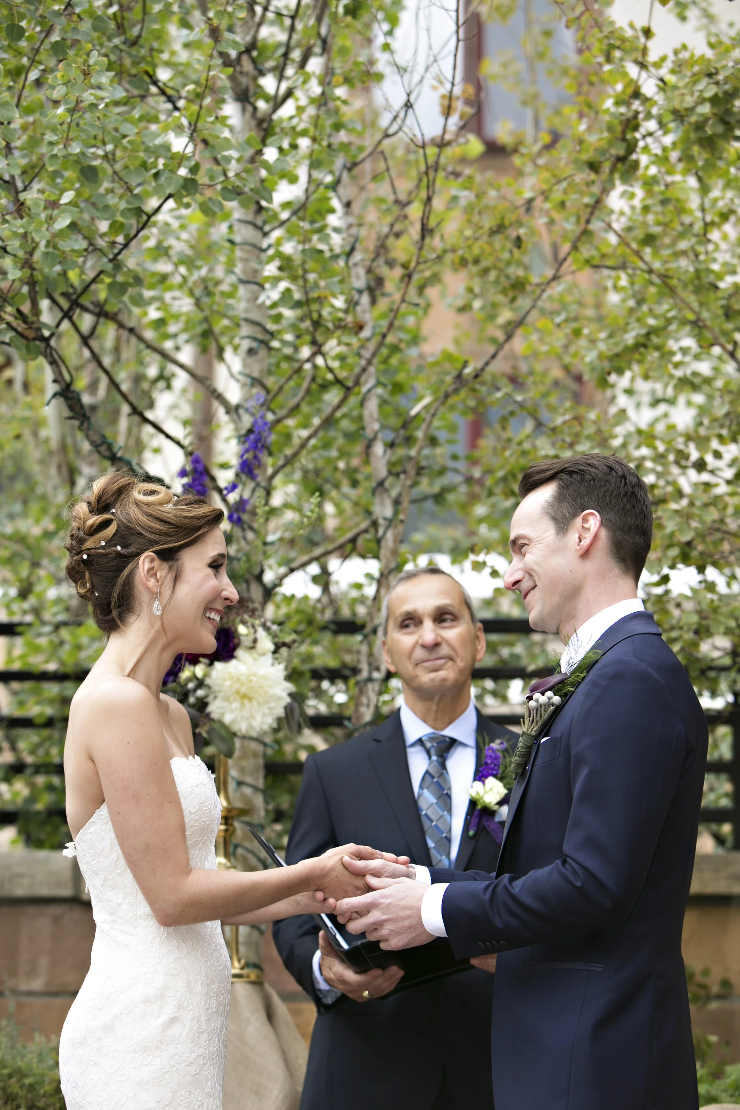 A wedding ceremony with a bride and groom holding hands and smiling at each other, with an officiant standing between them, outdoors surrounded by trees and greenery.