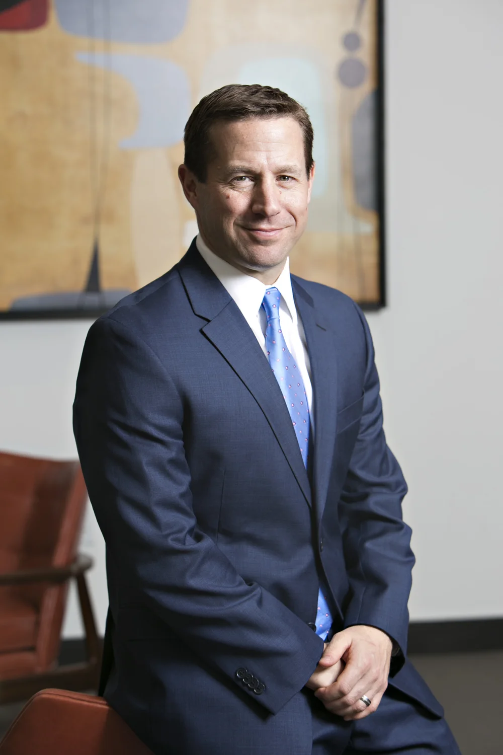 A man in a navy blue suit with a white shirt and a light blue tie with pink dots, sitting on a brown chair, smiling, with a colorful abstract painting on the wall behind him.
