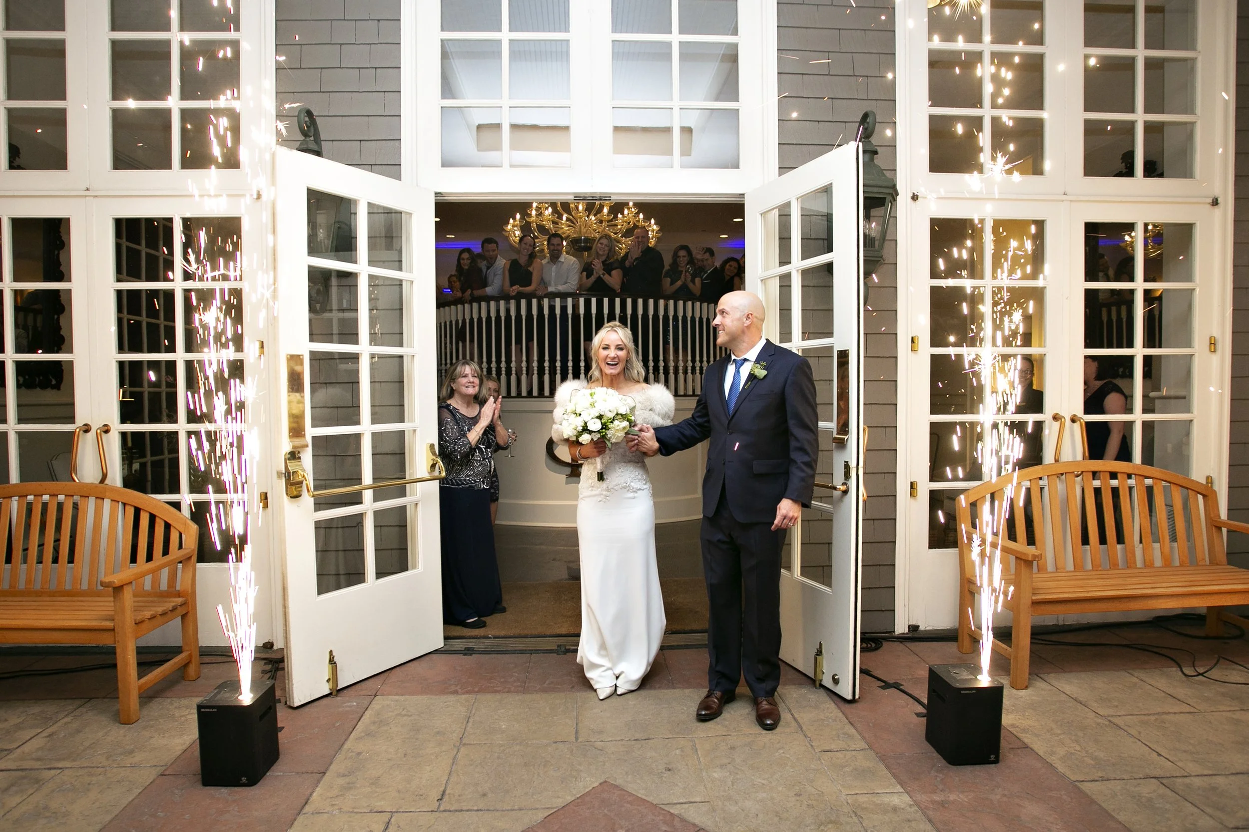 A bride and groom exit a building through open double doors, surrounded by sparklers, with onlookers cheering from inside and outside.