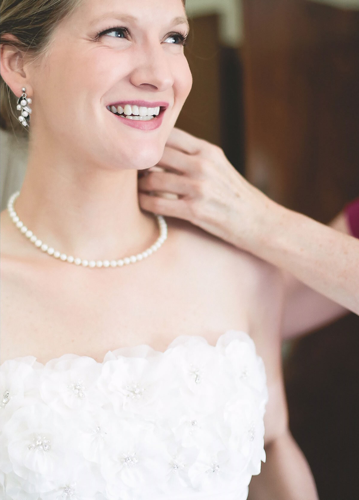  Smiling bride wearing custom designed pearl earrings and necklace, hand adjusting them on her ears 