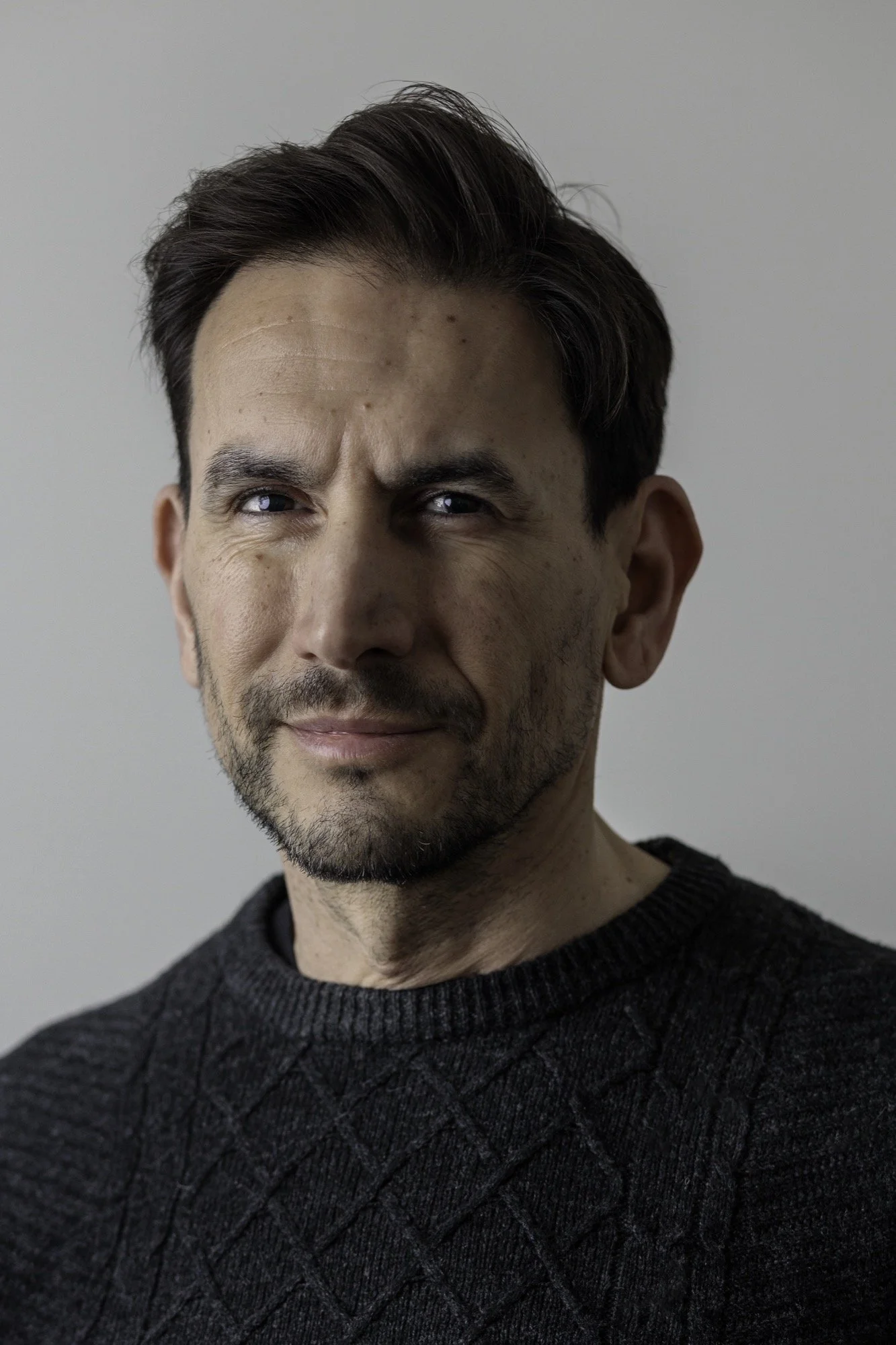 Close-up portrait of a man with dark hair, blue eyes, and facial hair, wearing a dark knitted sweater, against a plain light background.