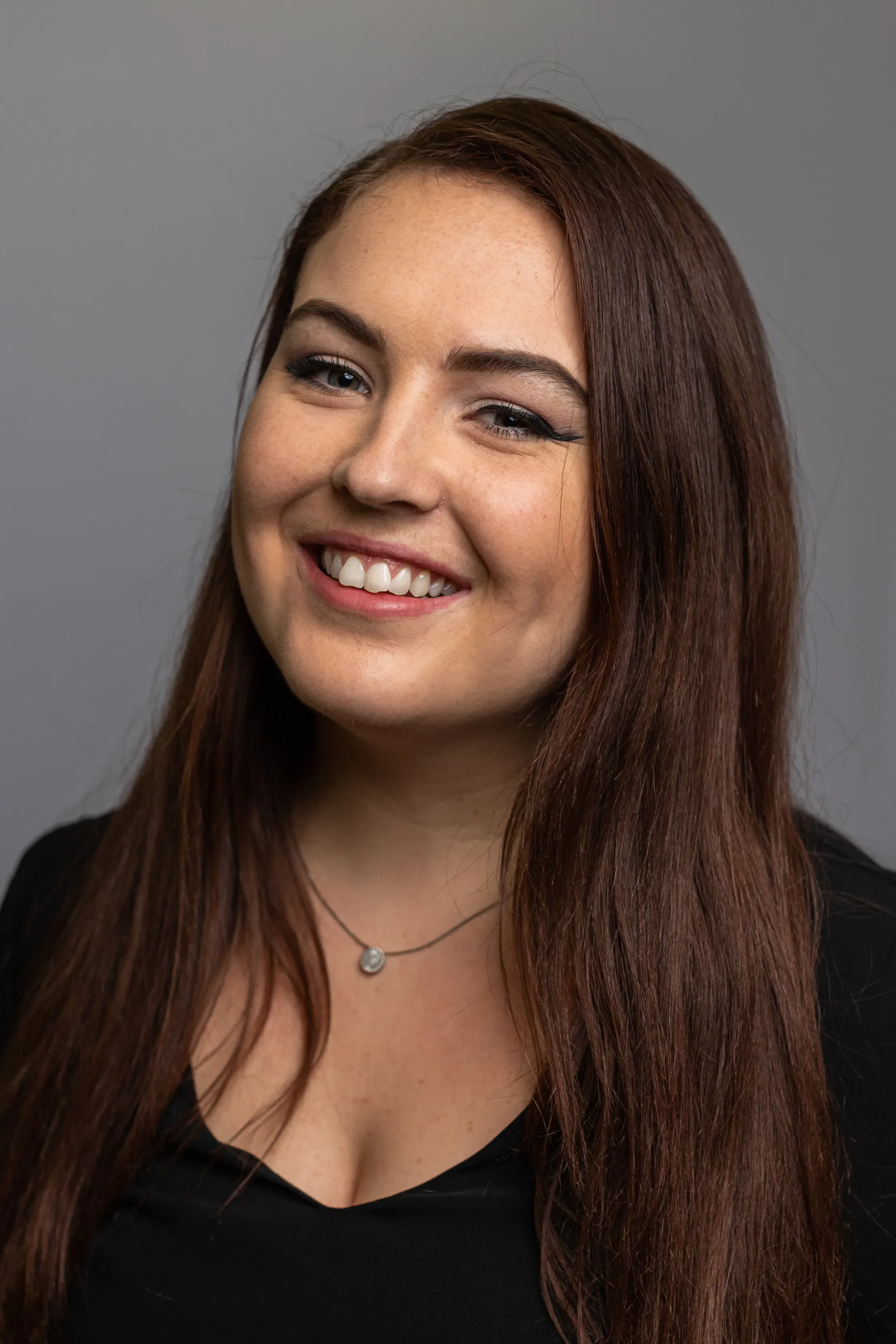 A young woman with long, wavy brown hair, smiling broadly, wearing a black top and a silver necklace, standing against a plain gray background.