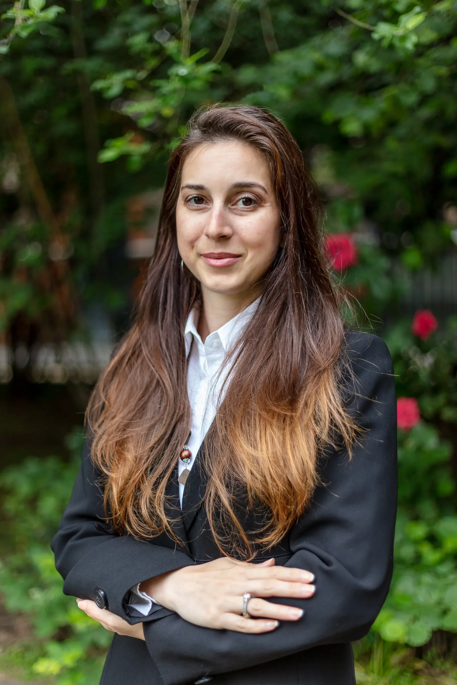 A woman with long brown hair and light skin, dressed in a black blazer and white blouse, standing outdoors with green trees and bushes in the background, arms crossed, smiling gently.