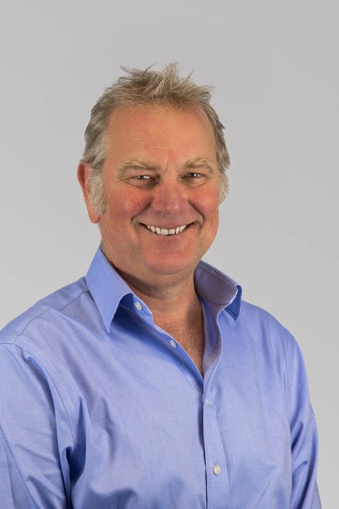 A middle-aged man with light brown hair smiling, wearing a blue button-up shirt, against a plain gray background.