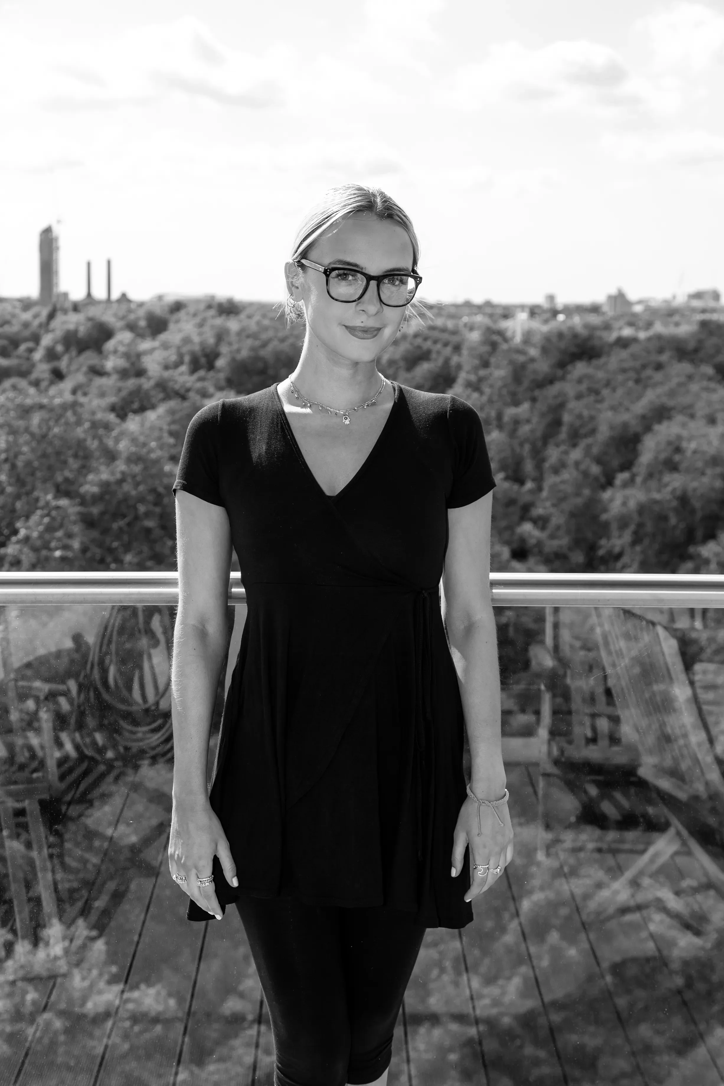 A woman with short hair, glasses, and jewelry, standing outdoors on a bridge with a cityscape and trees in the background, in black and white.