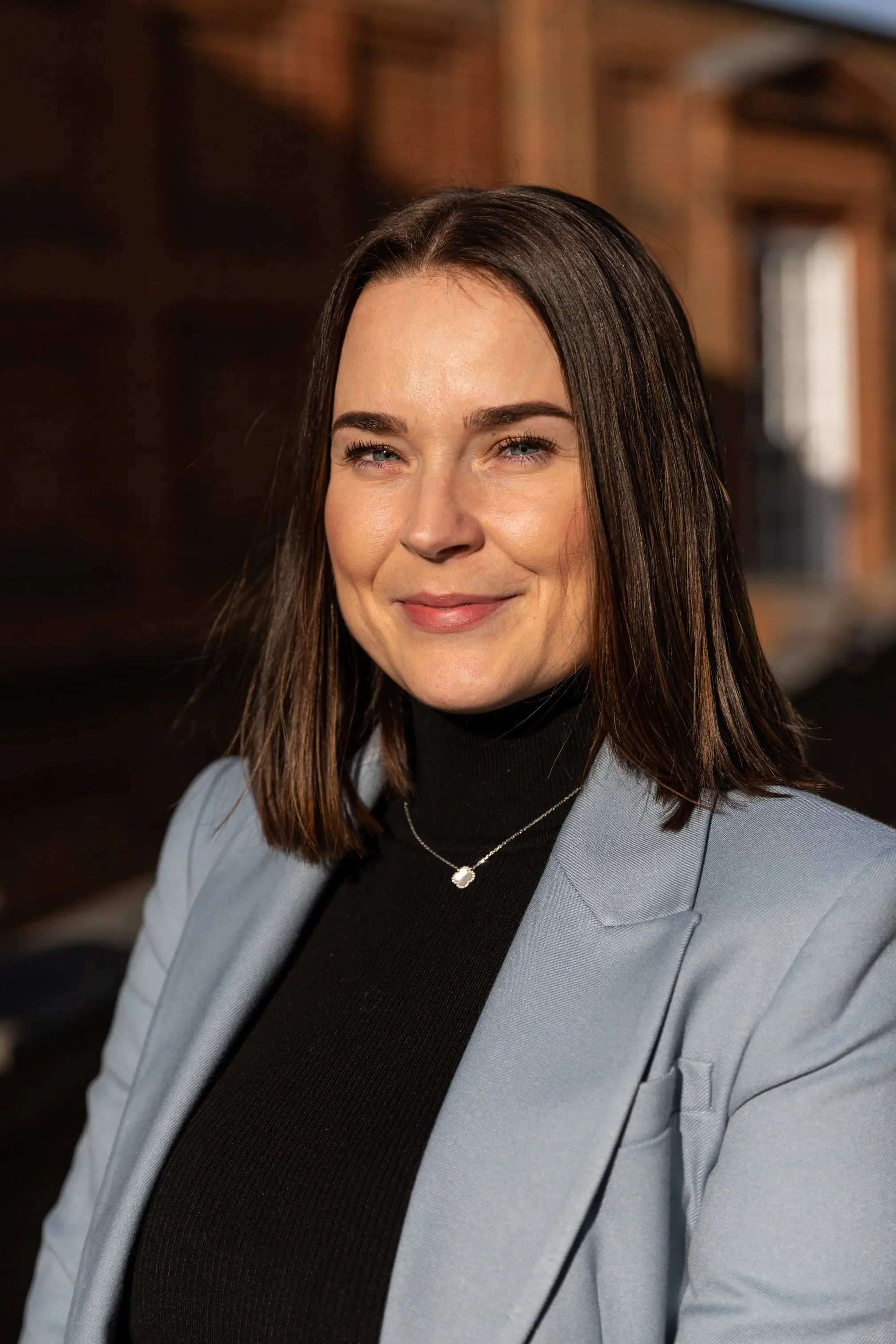 A woman with long brown hair and blue eyes, smiling, wearing a black turtleneck, a light blue blazer, and a silver necklace with a round pendant, standing outdoors with buildings in the background.