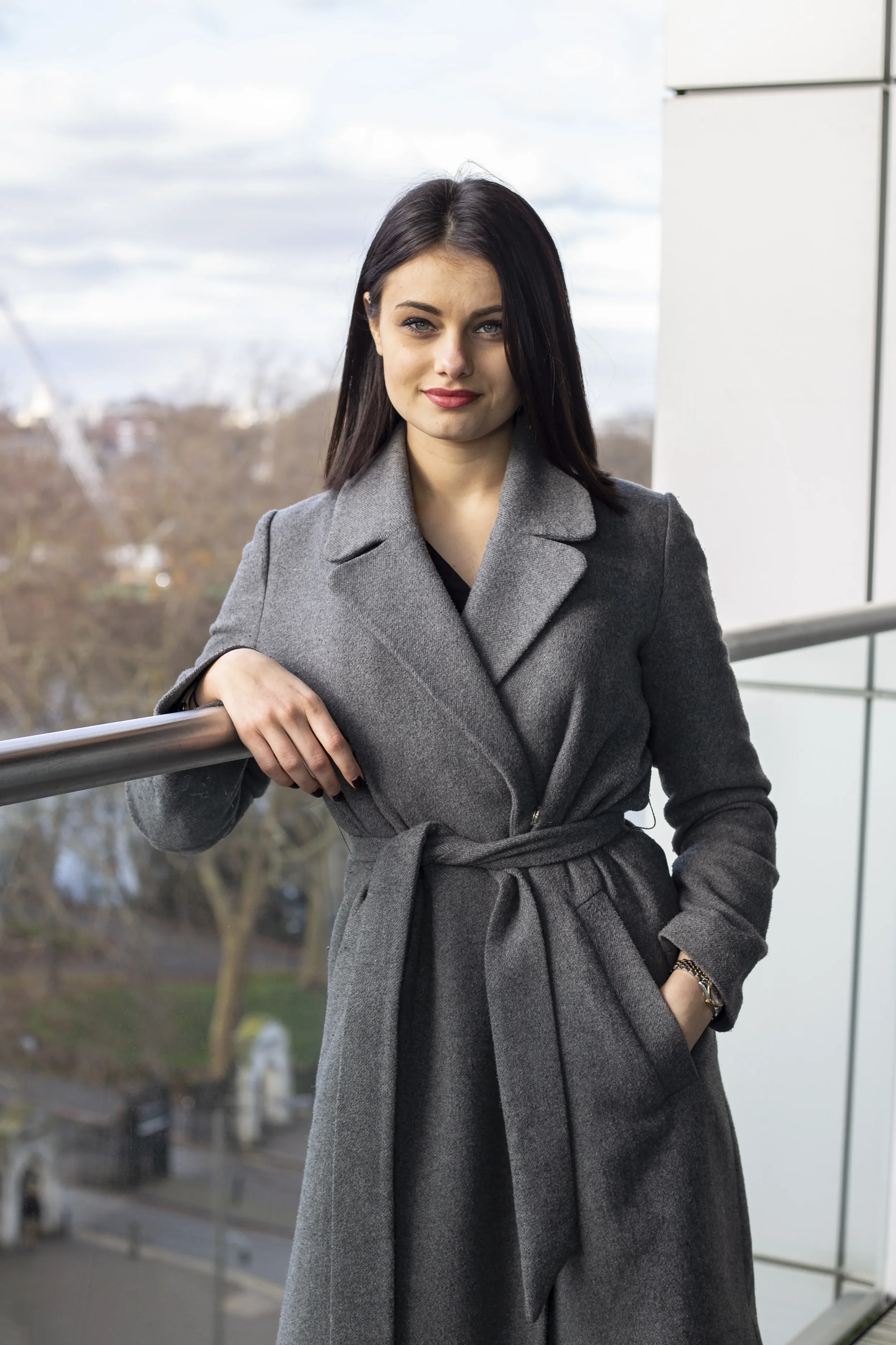 A woman with dark hair and fair skin wearing a gray coat standing outdoors on a balcony with urban trees and buildings in the background.