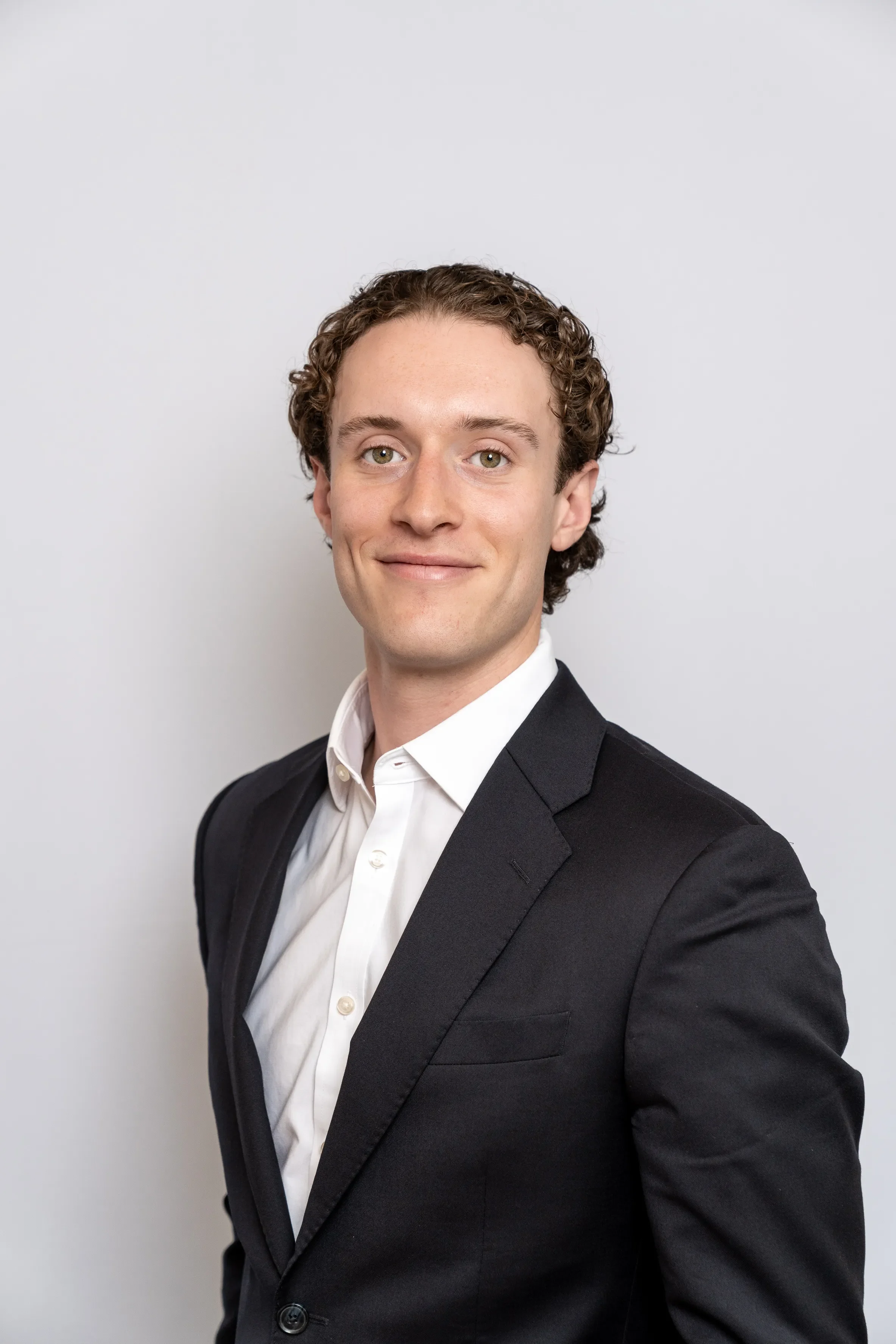 A portrait of a young man with curly brown hair, wearing a black suit jacket and a white dress shirt, standing against a plain white background.