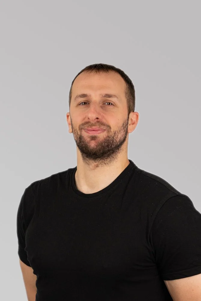 A man with short brown hair and a beard, wearing a black t-shirt, standing against a light gray background.