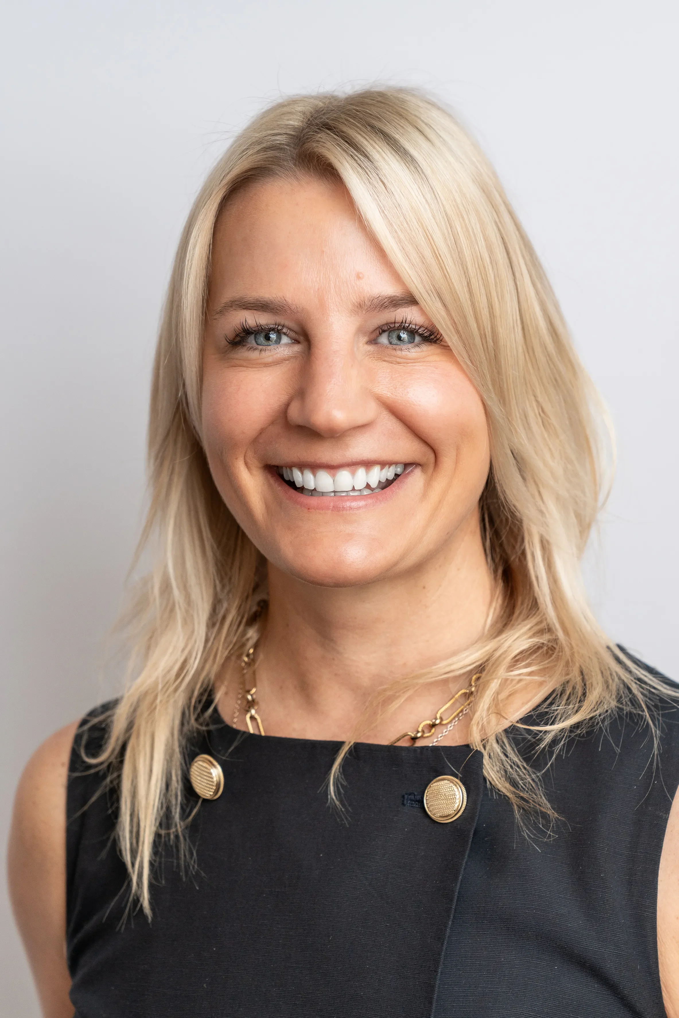Close-up of a smiling woman with blonde hair, wearing a black sleeveless top and gold jewelry, against a plain light background.