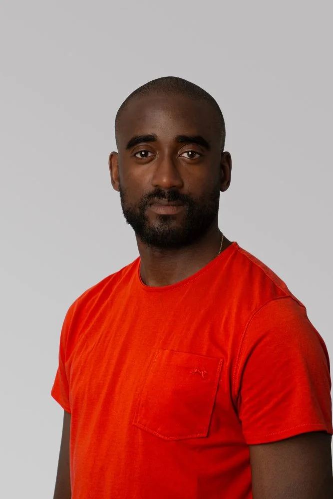 A man with dark skin, short hair, and a beard wearing an orange t-shirt against a plain gray background.