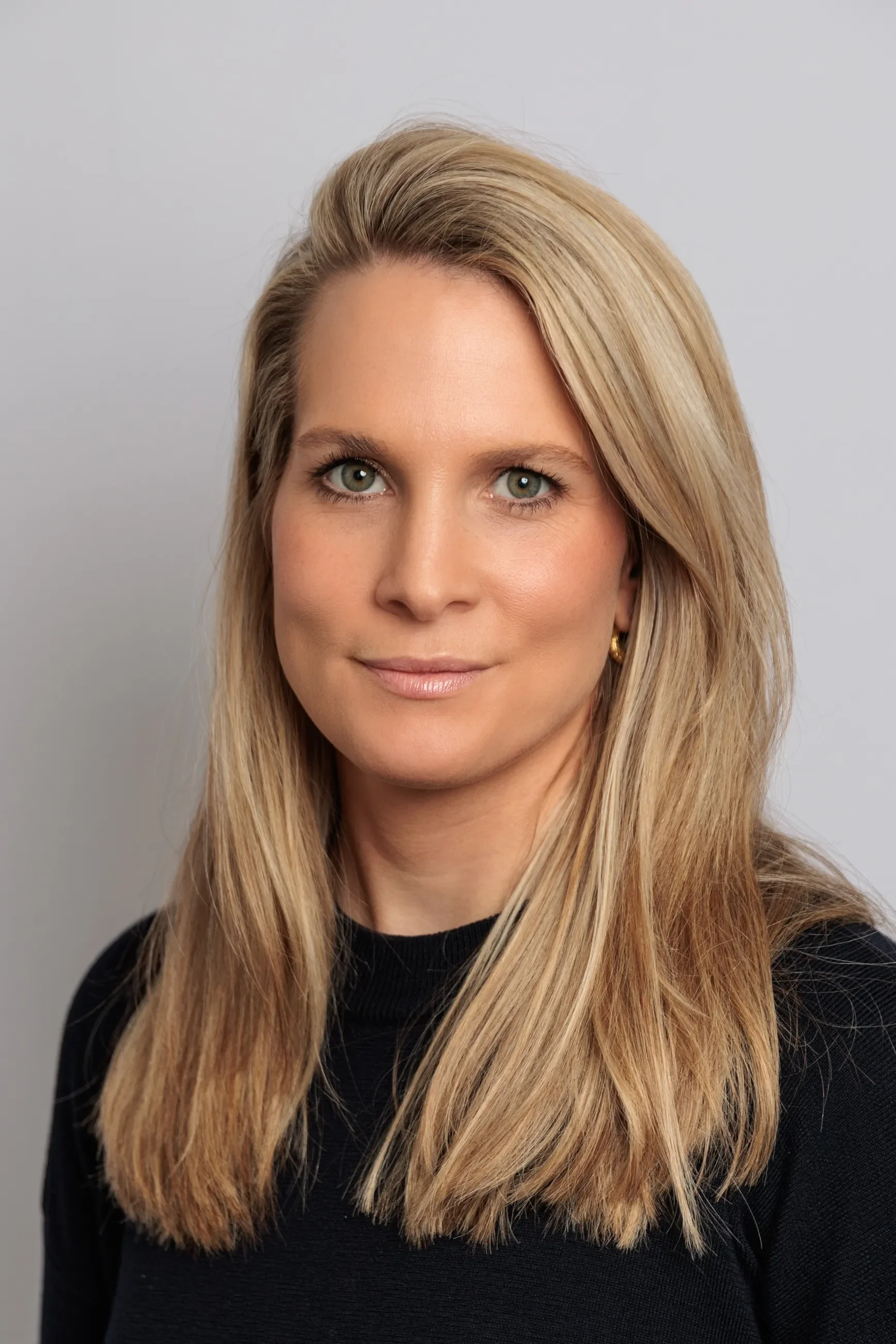 Close-up of a woman with long blonde hair, blue eyes, wearing a black top, smiling subtly, against a neutral background.
