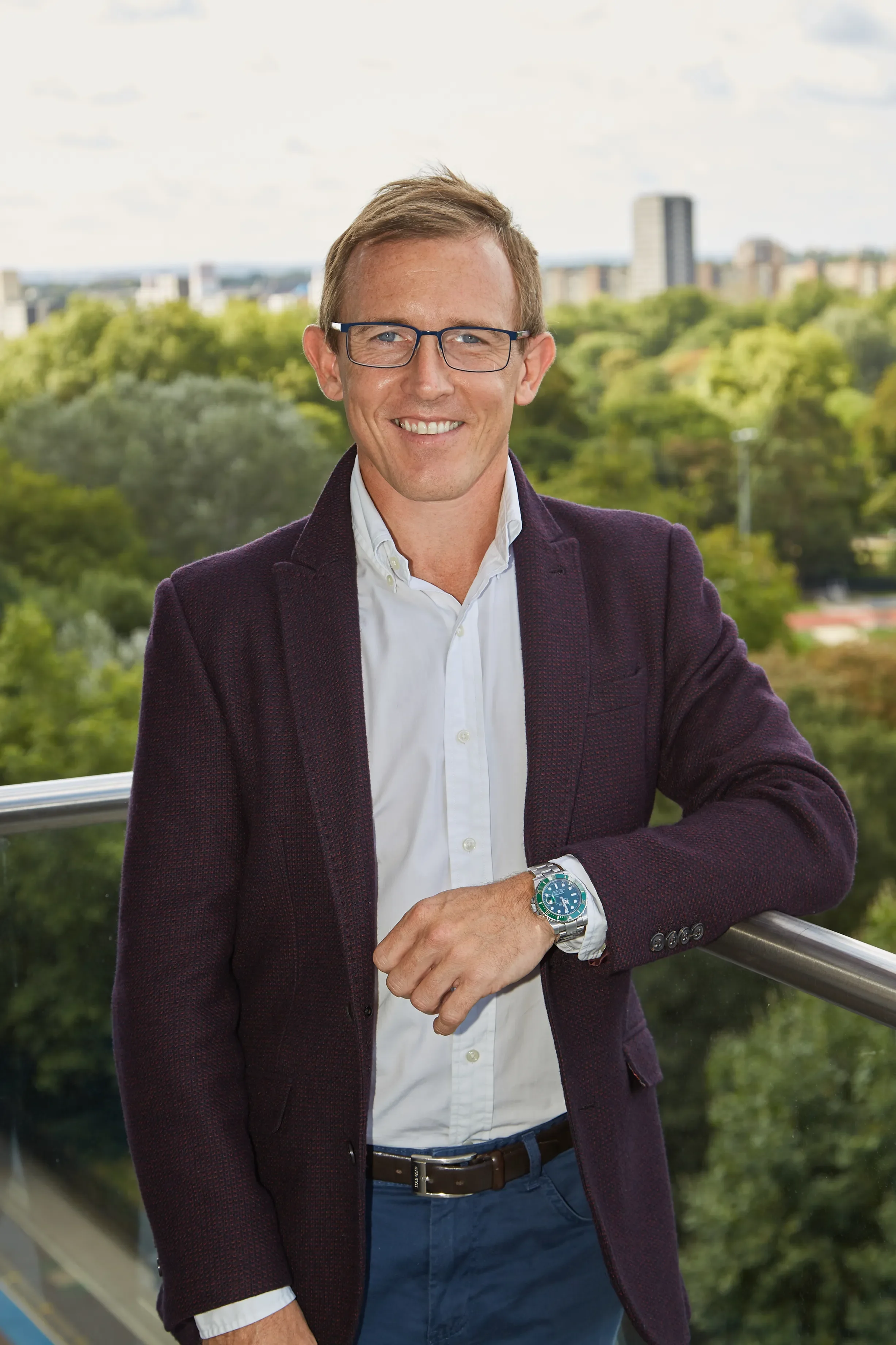 A man wearing glasses, a white shirt, dark trousers, and a maroon blazer, standing outdoors on a balcony with a cityscape and green trees in the background.