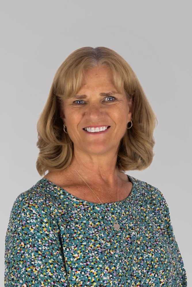 A smiling middle-aged woman with blonde hair wearing a colorful patterned blouse and silver jewelry against a plain light gray background.