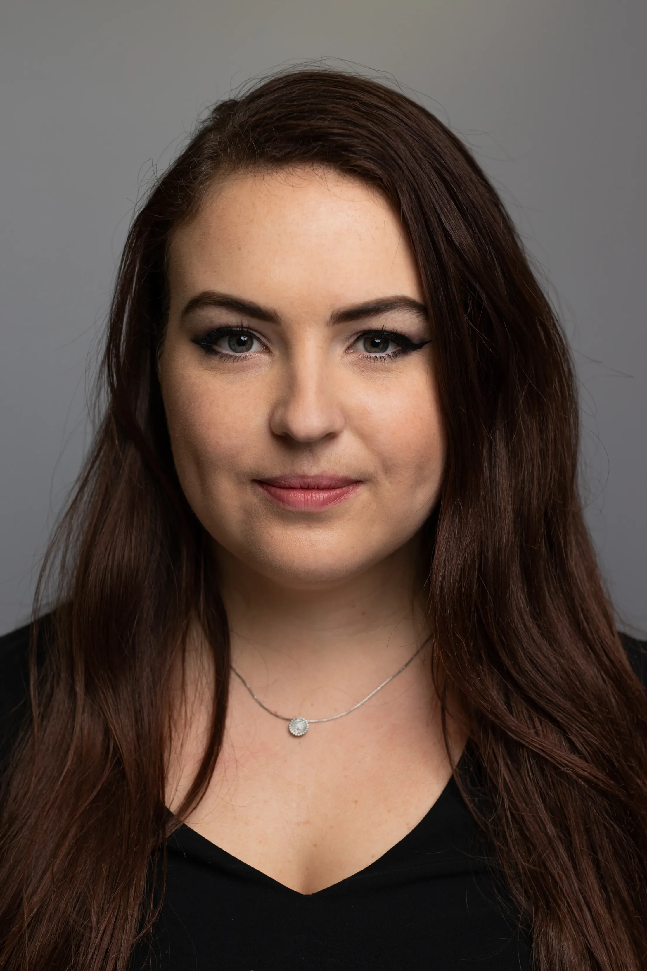 Close-up of a woman with long dark brown hair, blue eyes, and wearing makeup with winged eyeliner. She is wearing a black top and a silver necklace with a round pendant, against a plain gray background.