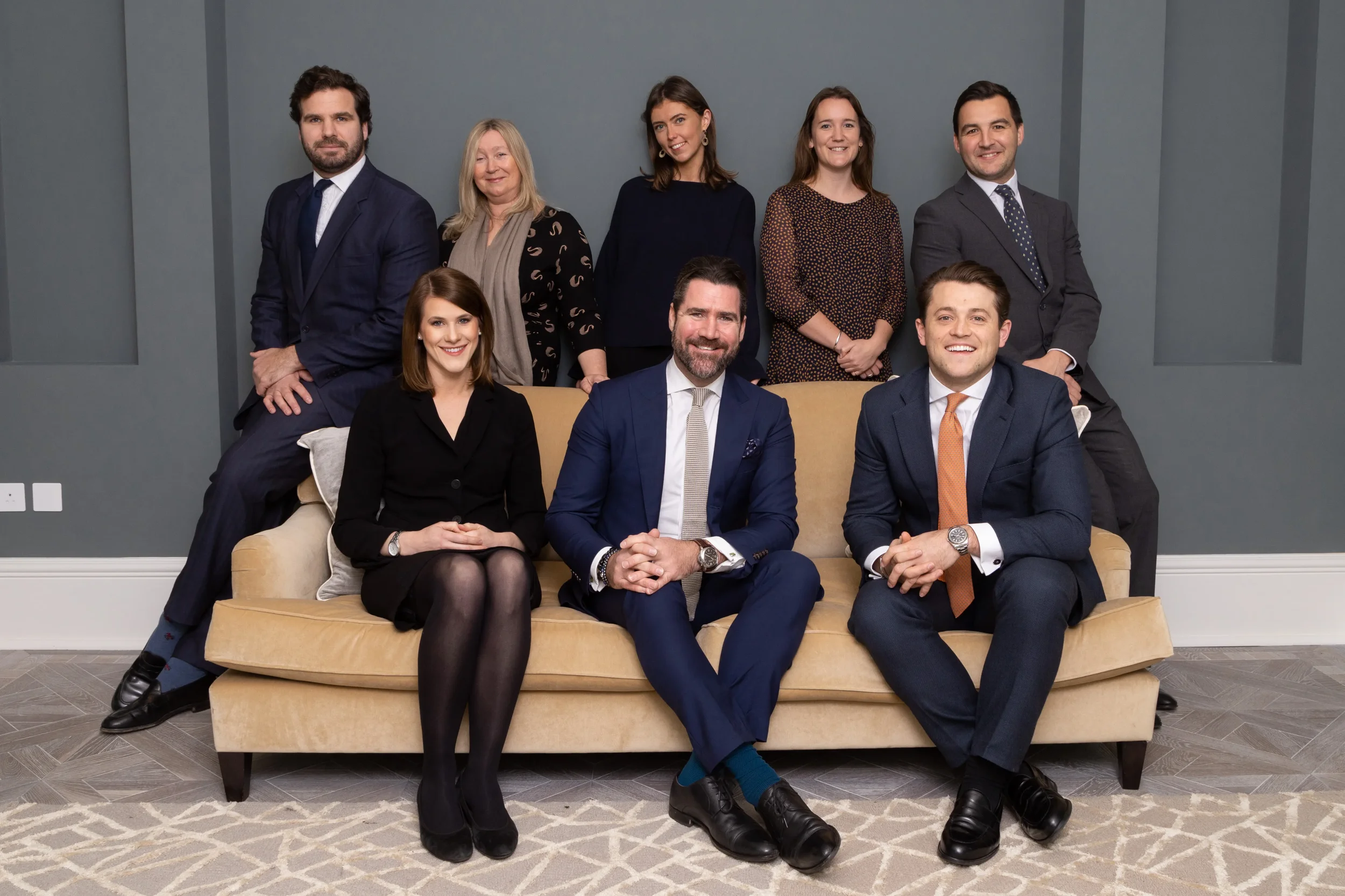 A group of nine diverse professionally dressed people gathered in a modern living room. They are smiling and posed for a formal group photo, with some seated on a beige sofa and others standing behind. The background features a gray wall and a patterned gray rug on the floor.