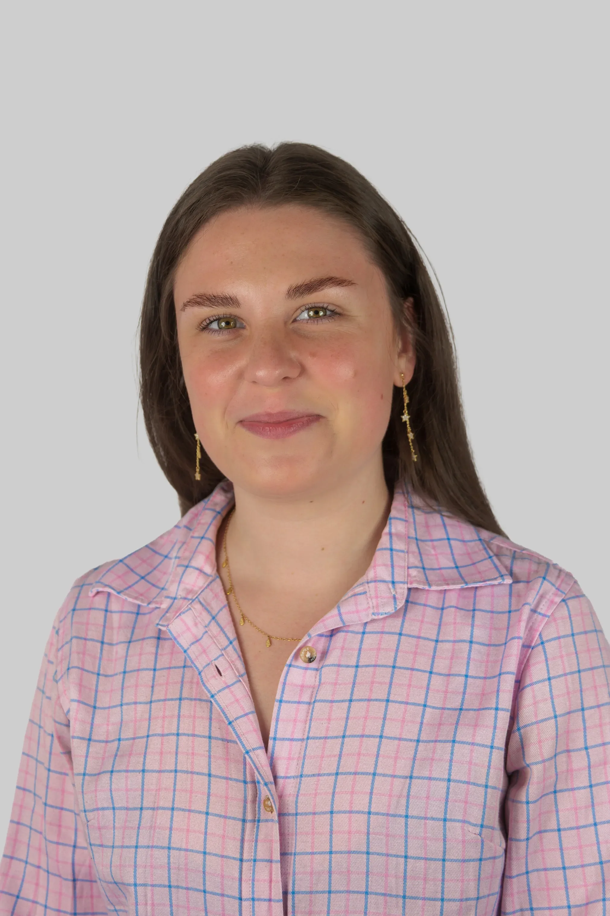 Close-up of a young woman with brown hair, green eyes, and earrings, wearing a pink and blue checkered shirt, smiling softly against a plain light gray background.