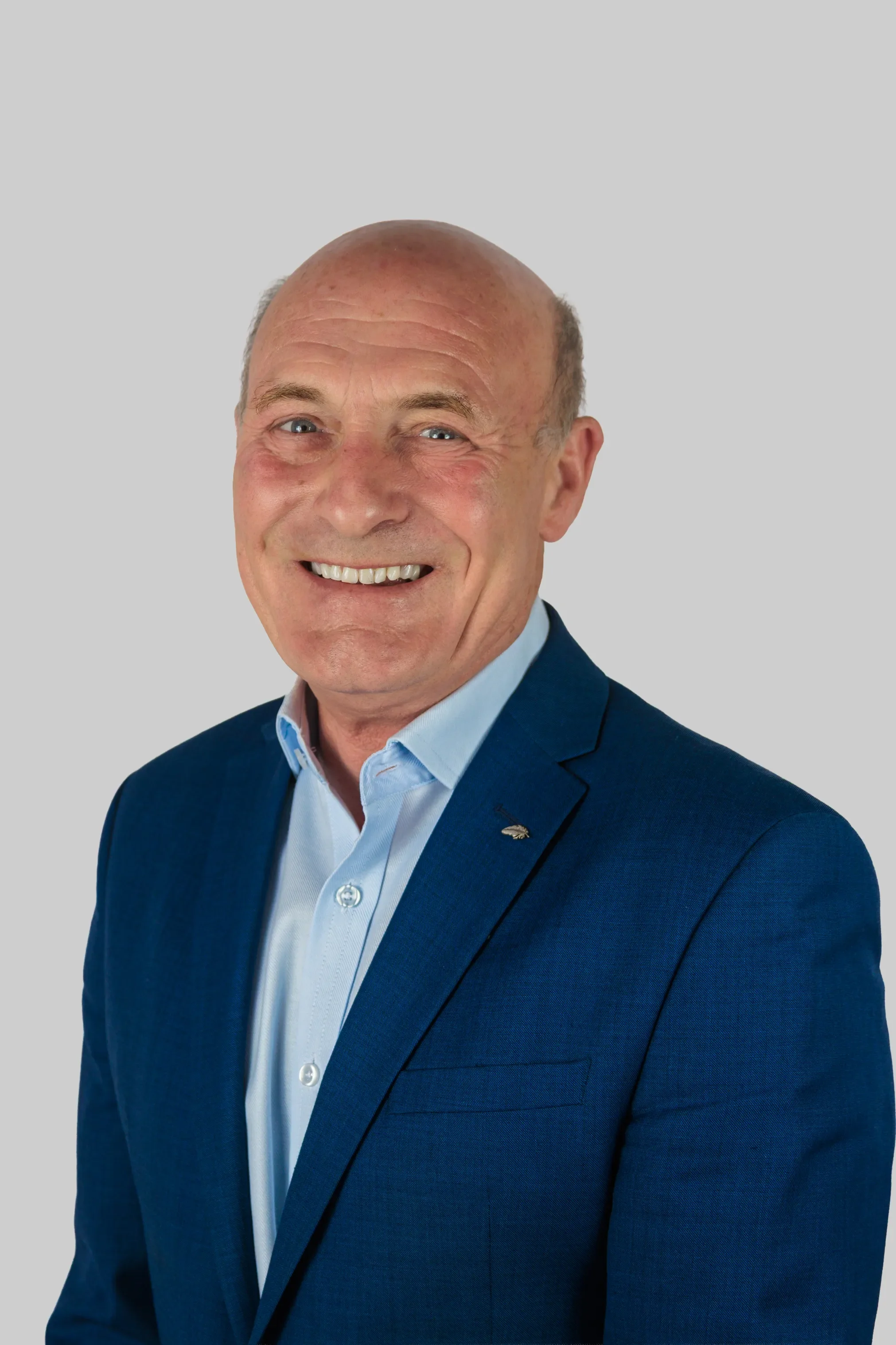 Portrait of a smiling middle-aged man with a bald head, wearing a blue suit and white shirt, against a plain gray background.
