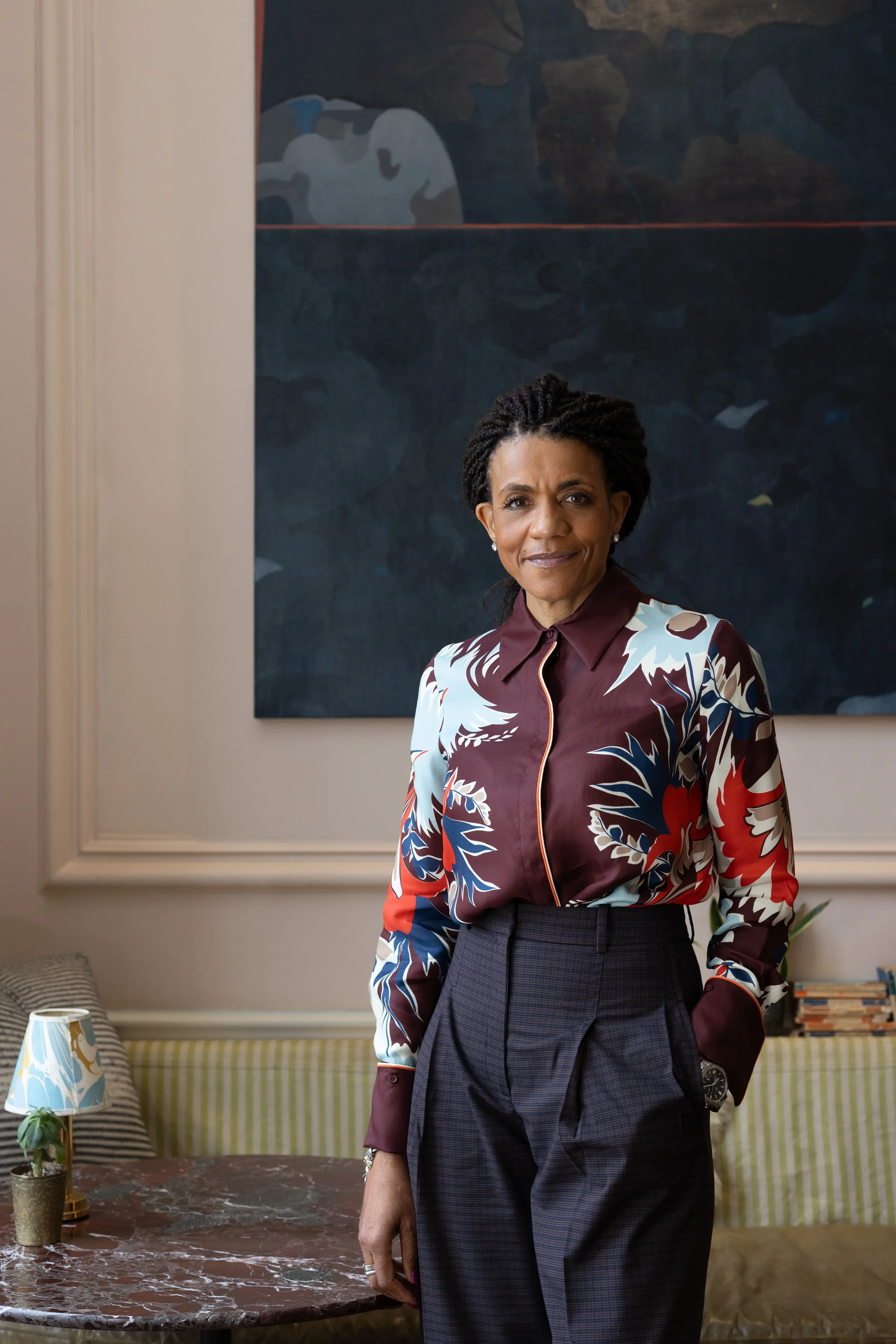 A woman standing in front of a large abstract painting, wearing a floral patterned blouse and dark plaid trousers, with a table and bookshelf in the background.