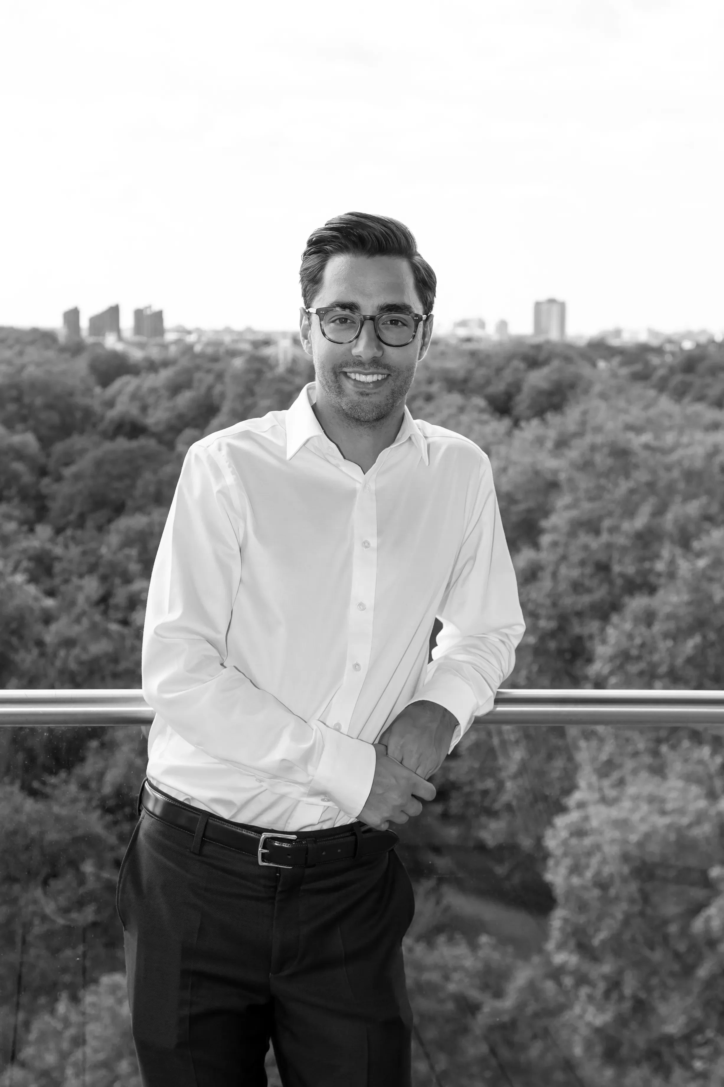 A professional man wearing glasses, a white shirt, and black pants standing outdoors with a cityscape and trees in the background.