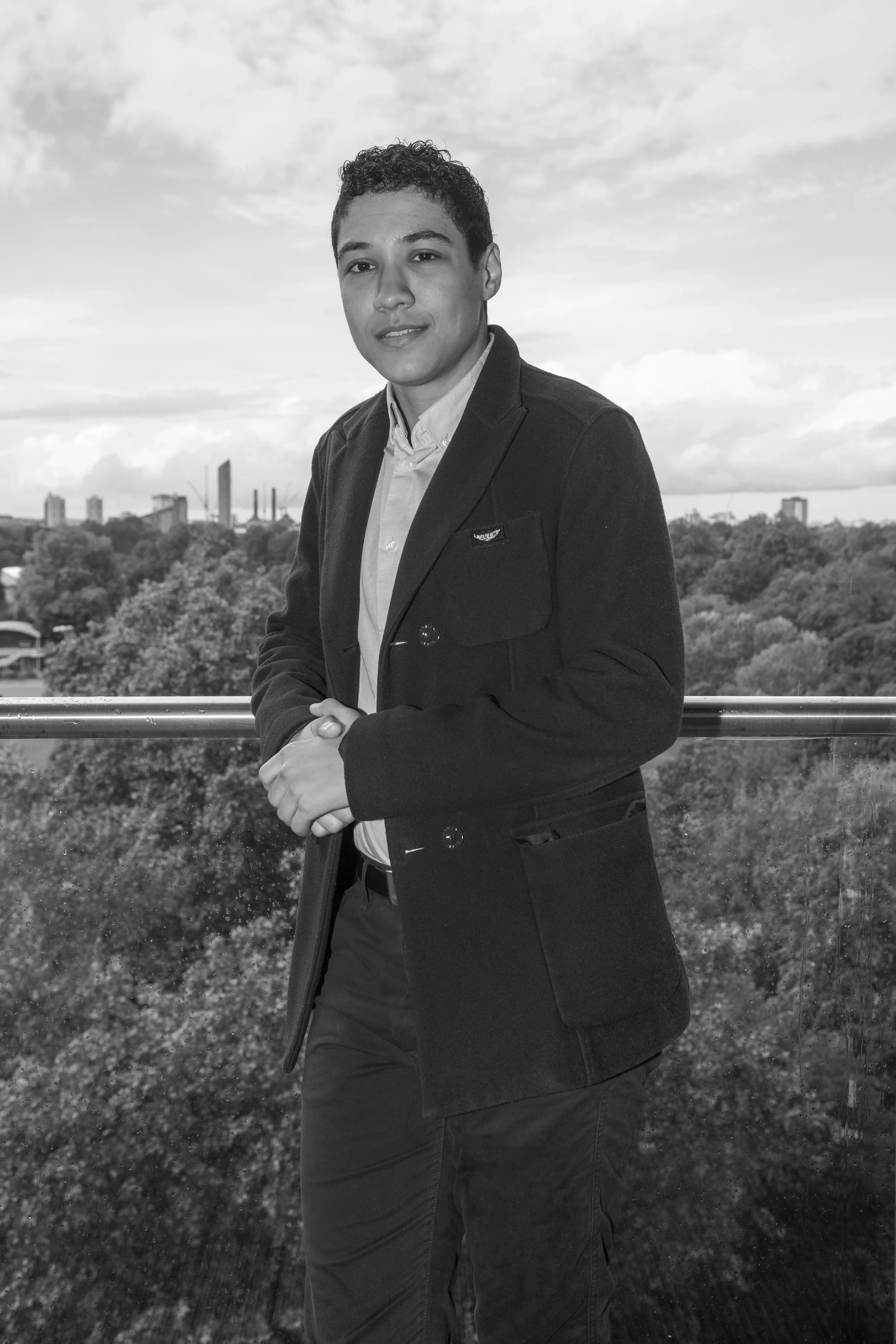 A young man with curly hair dressed in a blazer and shirt standing outdoors on a balcony with city skyline and cloudy sky in the background, in black and white.