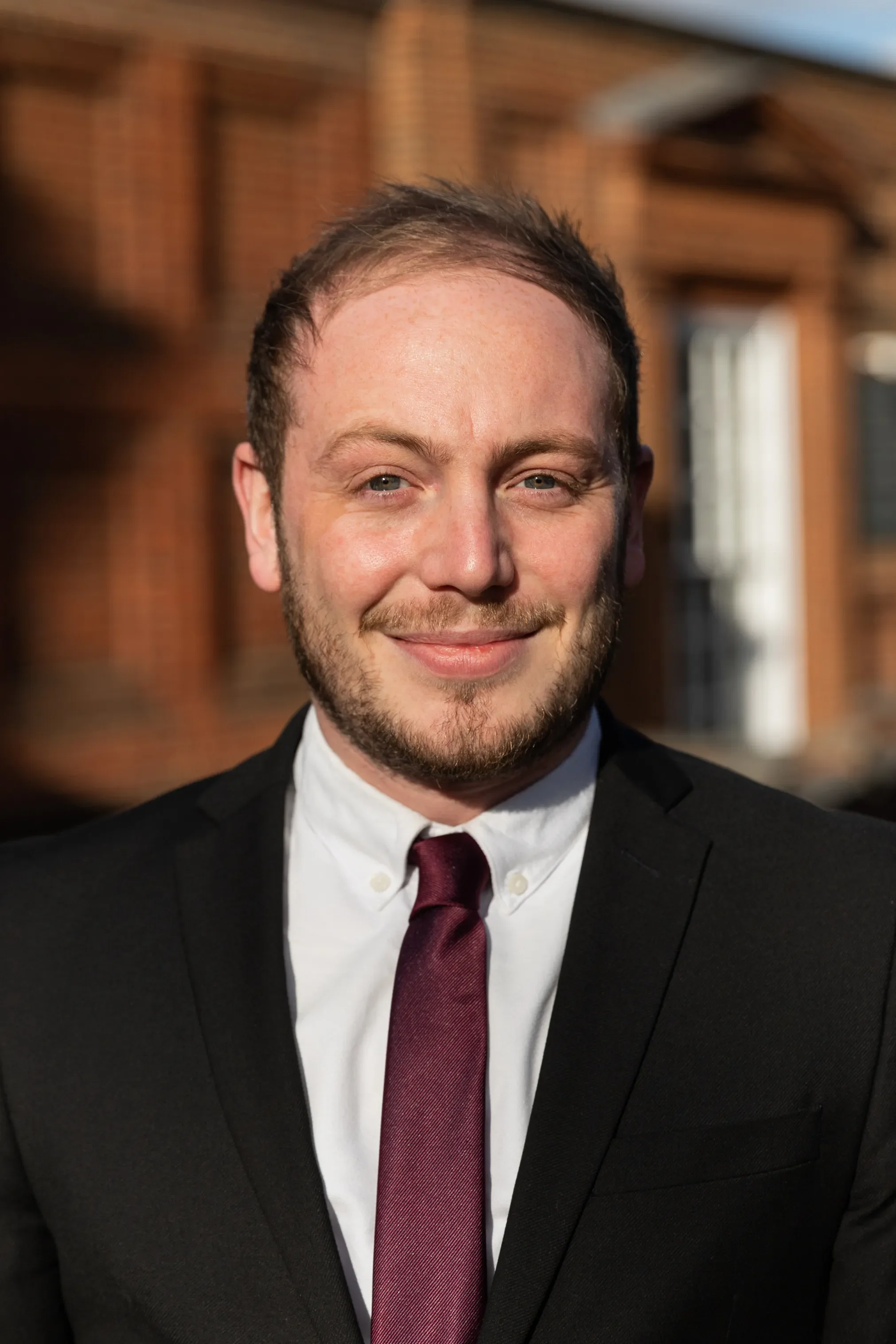 A smiling man in a black suit, white shirt, maroon tie, with a background of wooden building exterior