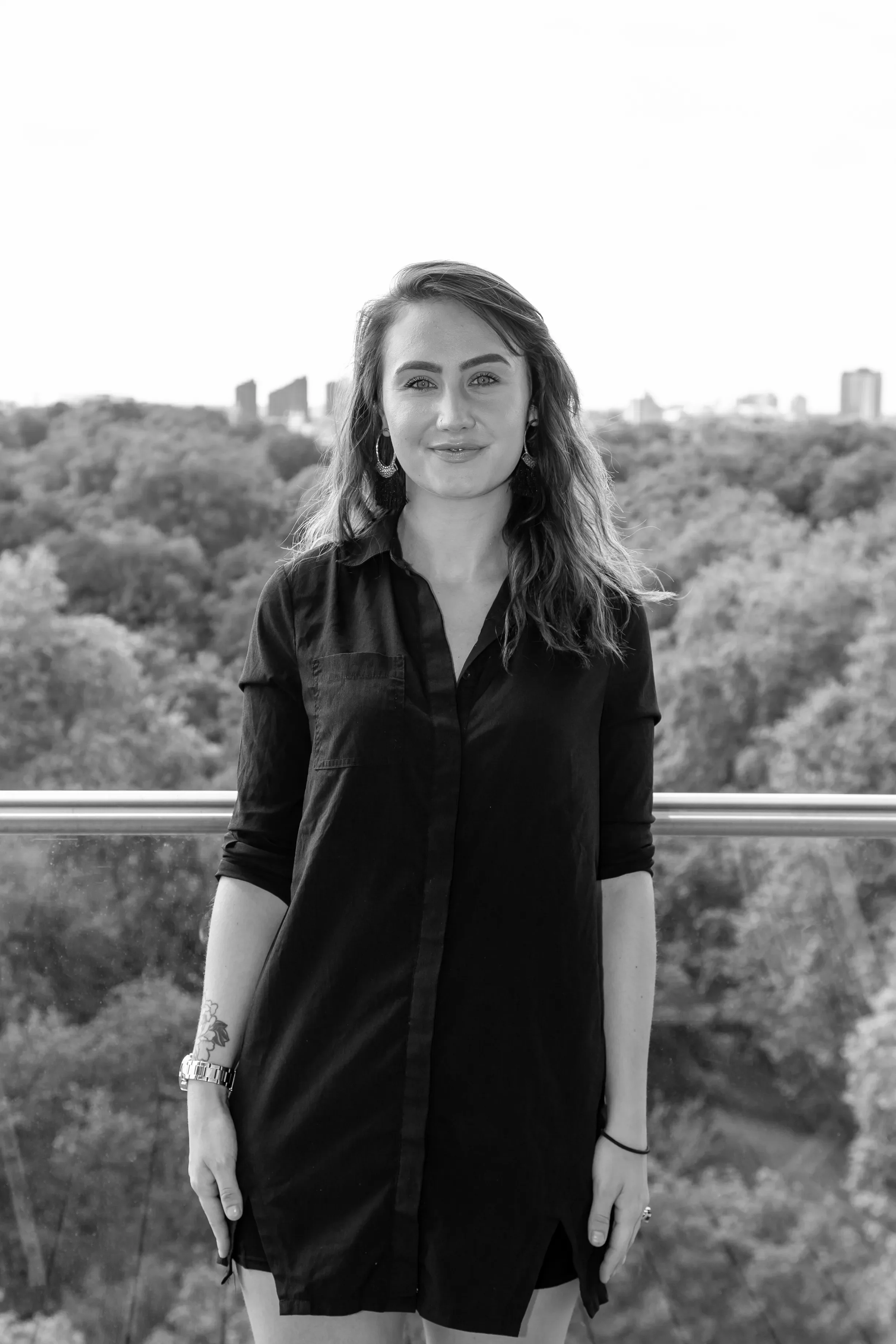 Black and white photo of a young woman with long, wavy hair, wearing a button-up shirt, standing outdoors with trees and city buildings in the background.