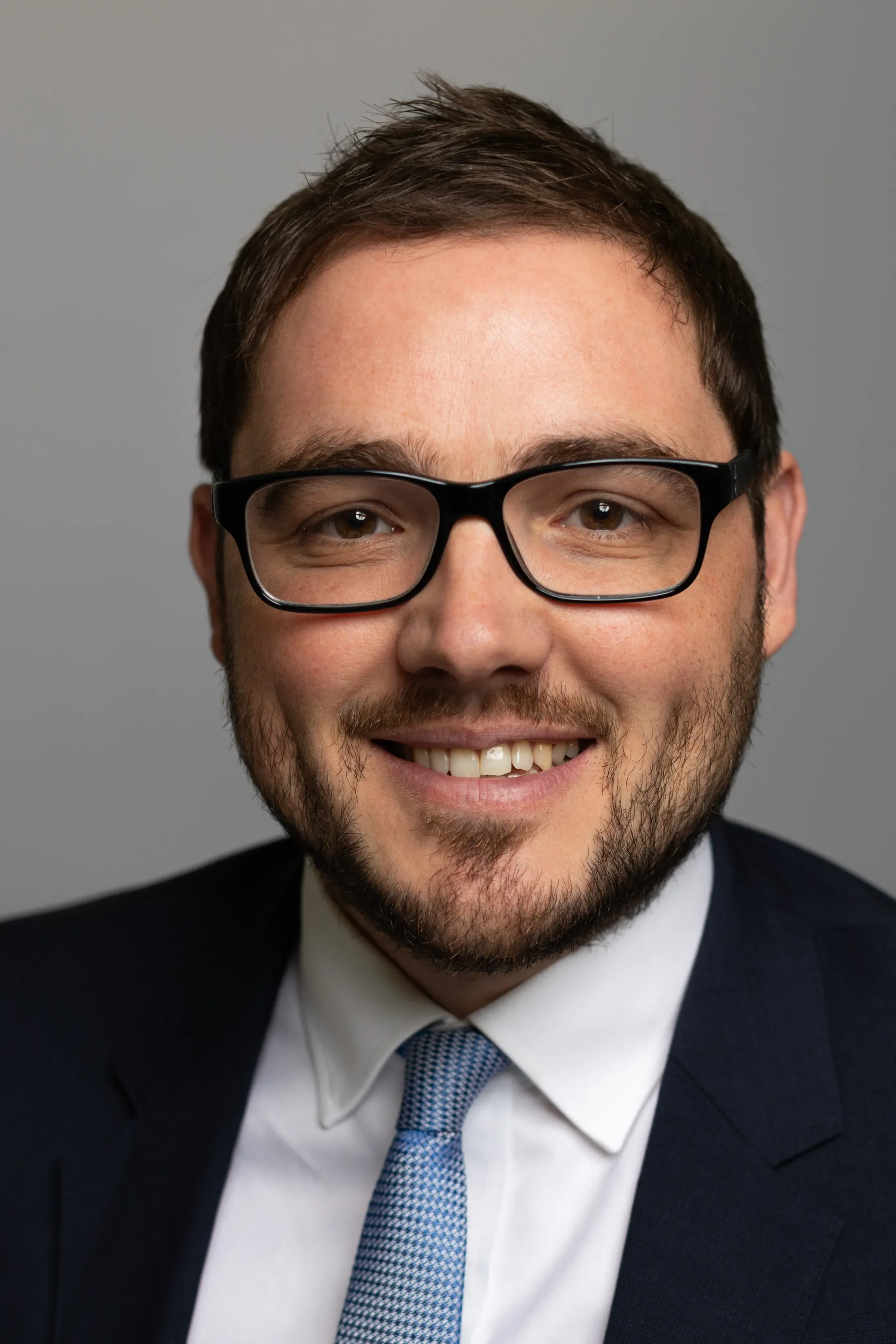 A smiling man with short brown hair, beard, and glasses, wearing a dark suit, white shirt, and blue tie, against a plain gray background.