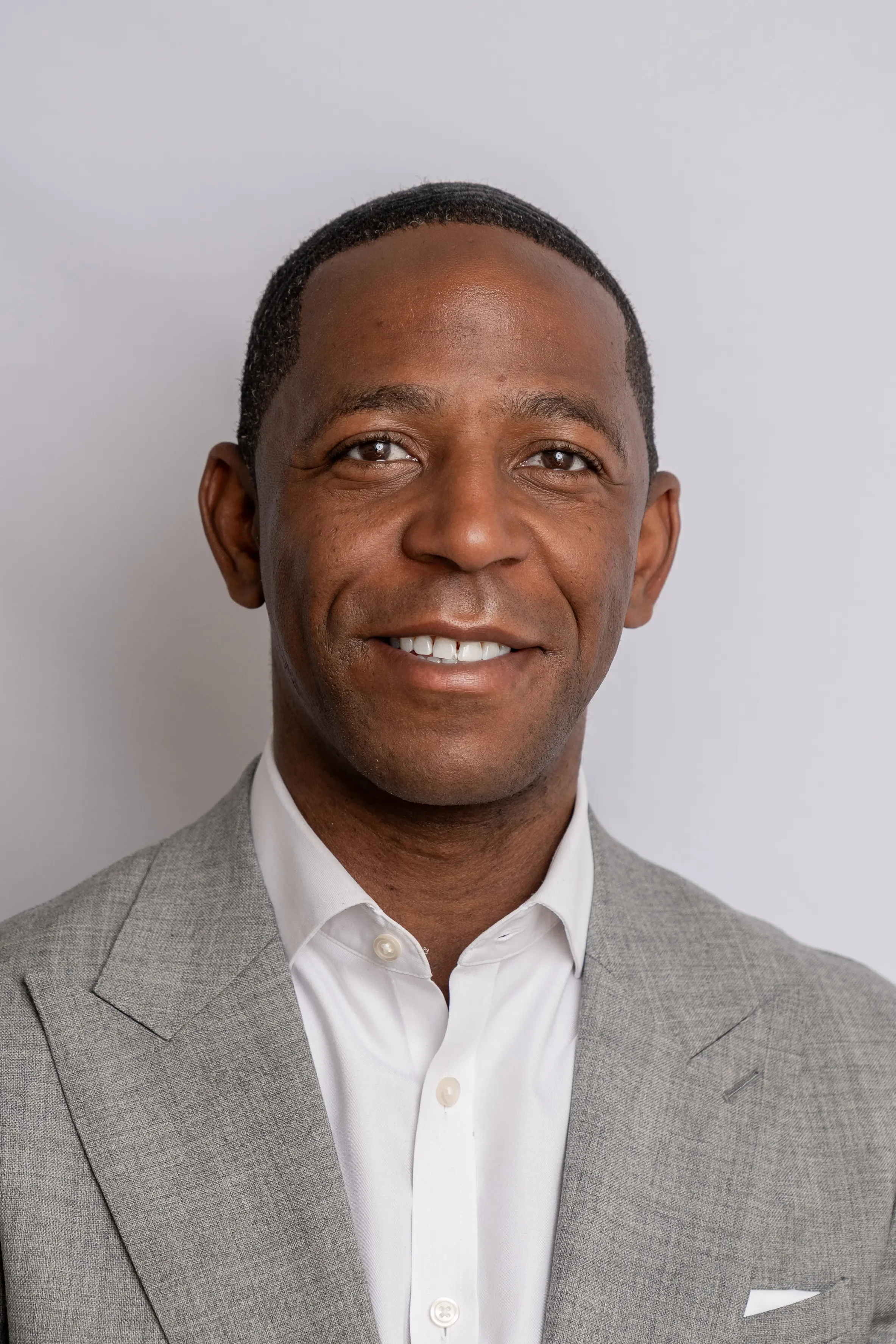 Close-up of a smiling man wearing a gray suit and white shirt against plain background.