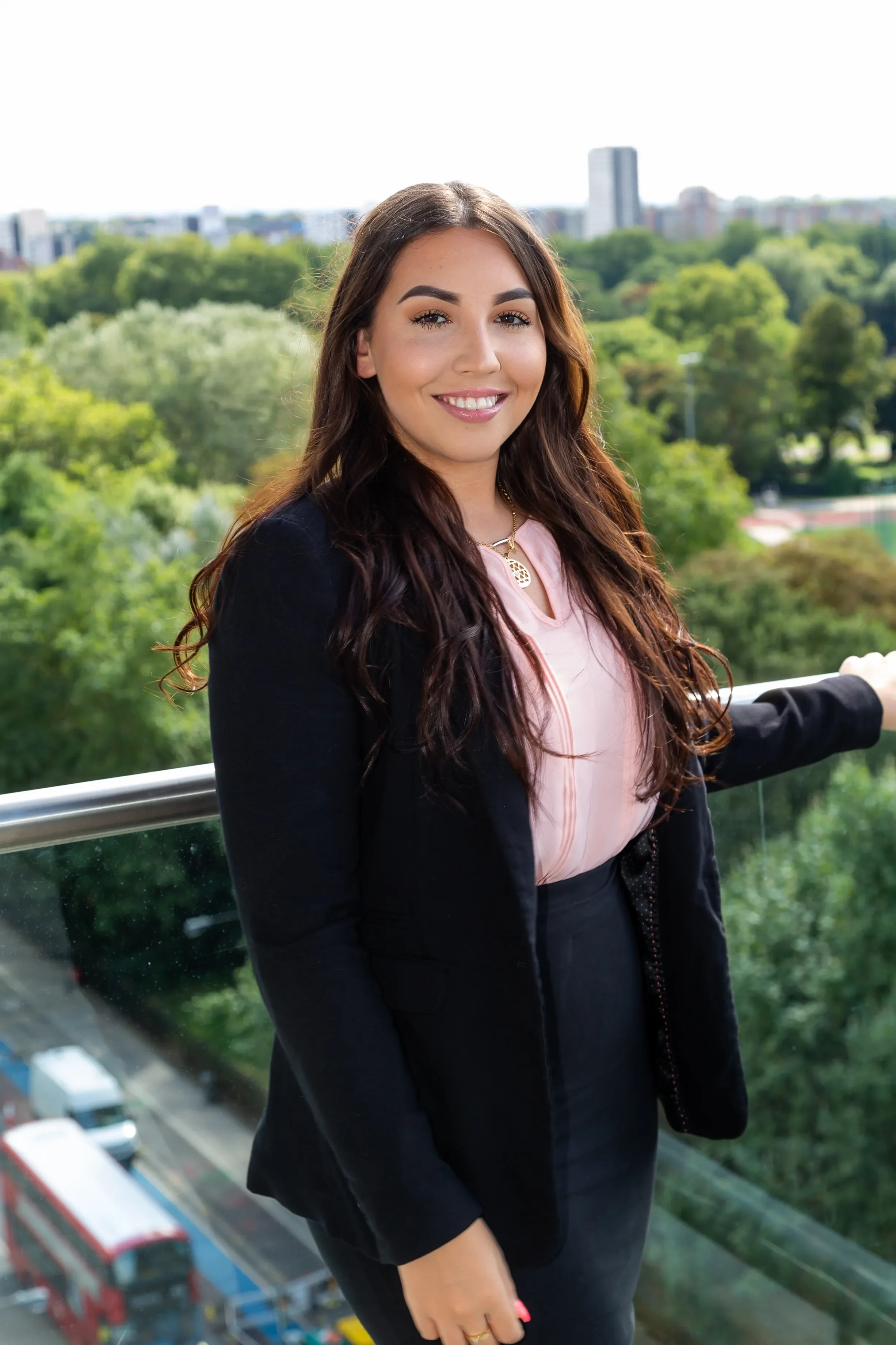 A woman with long brown hair, wearing a pink blouse and black blazer, standing on a balcony with a view of a city park, trees, and buildings in the background, smiling at the camera.