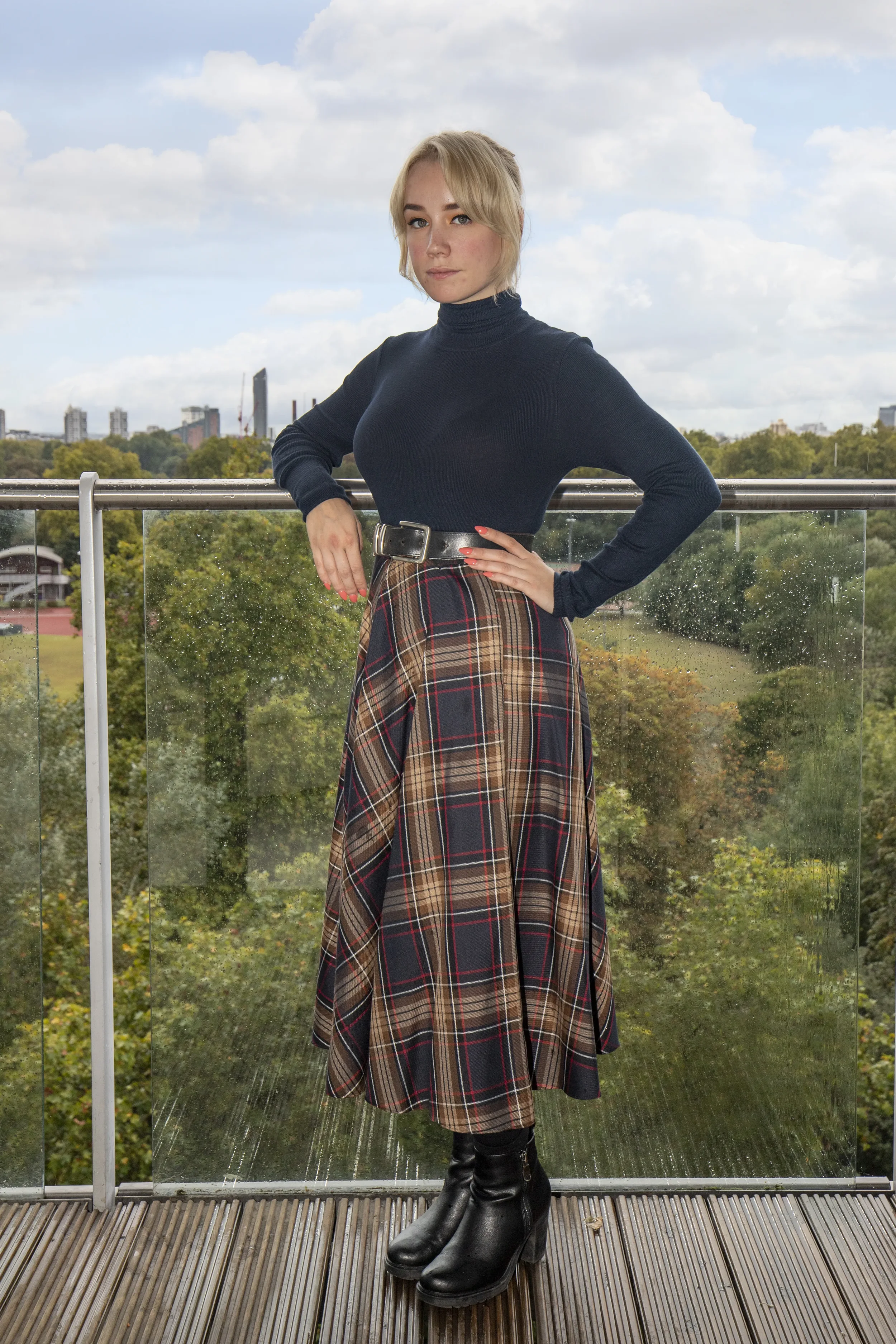 A woman standing on a balcony with a cityscape background, wearing a black turtleneck sweater, a plaid skirt, and black boots.