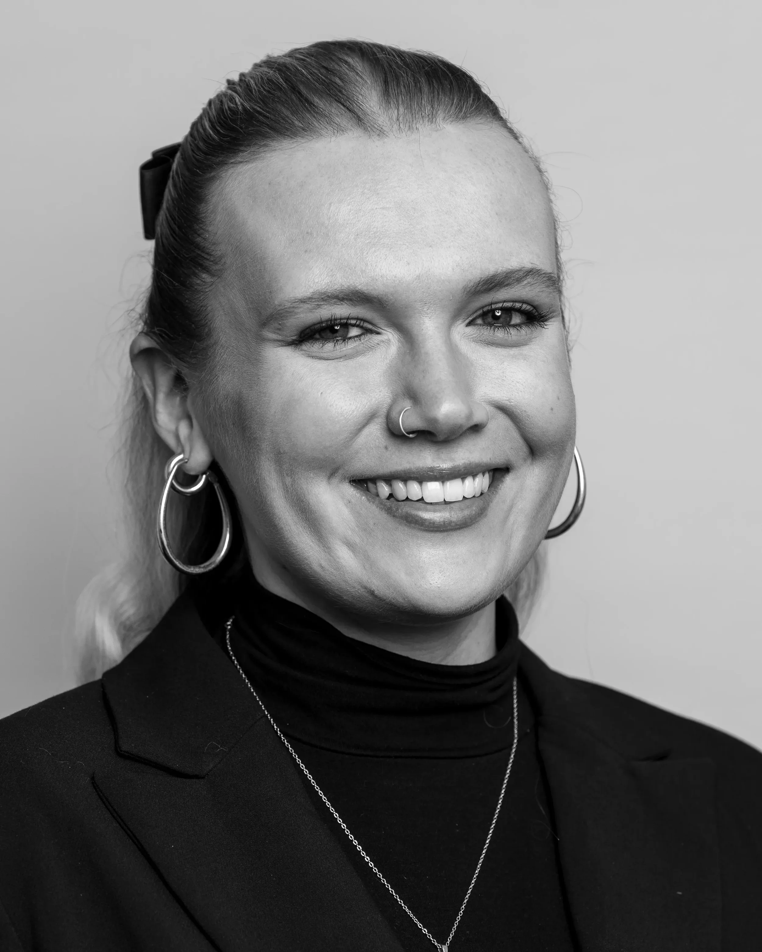 Close-up black and white photo of a smiling woman with hoop earrings, a nose ring, and a necklace, wearing a turtleneck and styled hair.