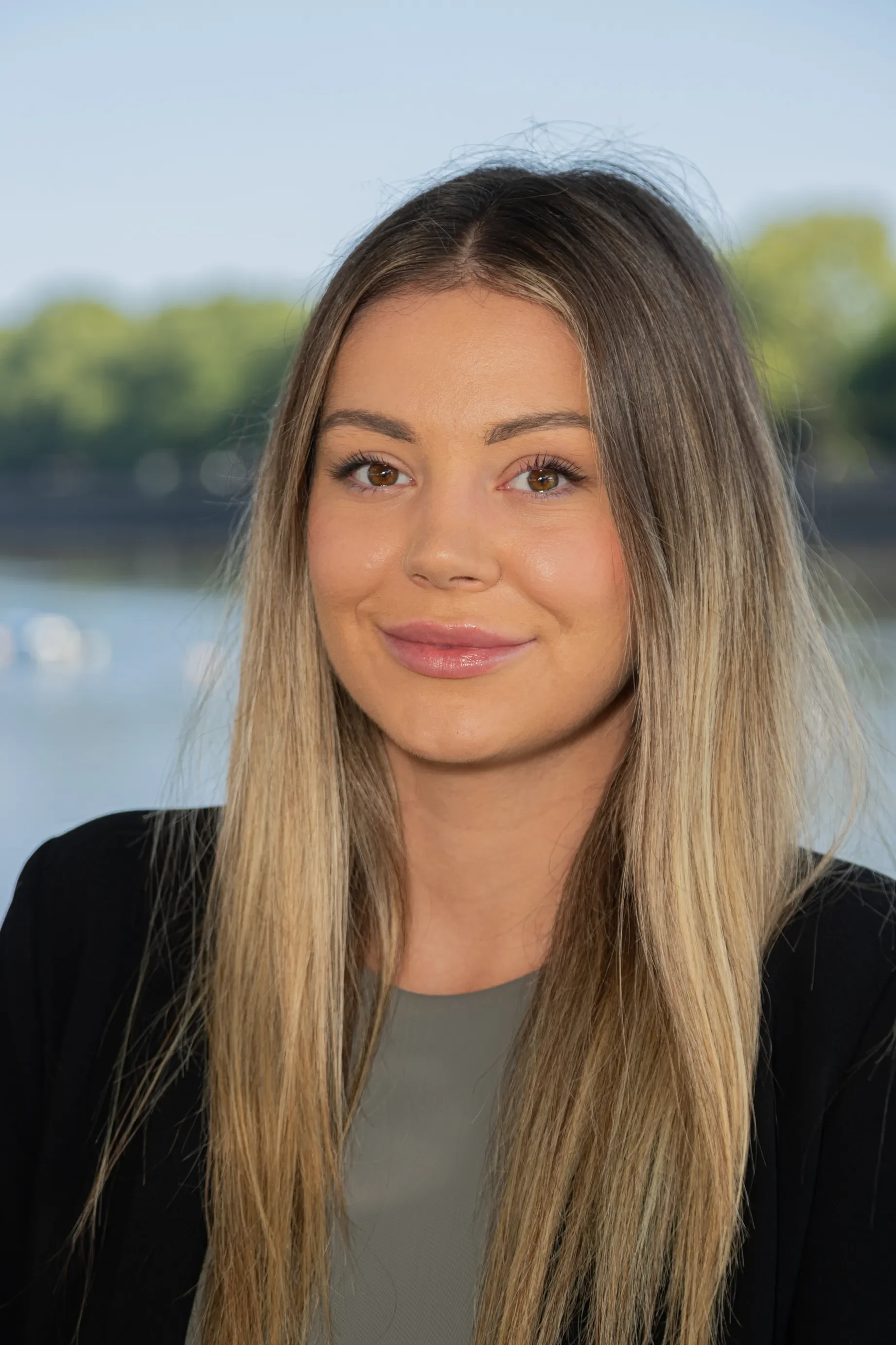 A young woman with long blonde hair, brown eyes, and light makeup, wearing a black blazer and a grey top, outdoors with blurred trees and water in the background.