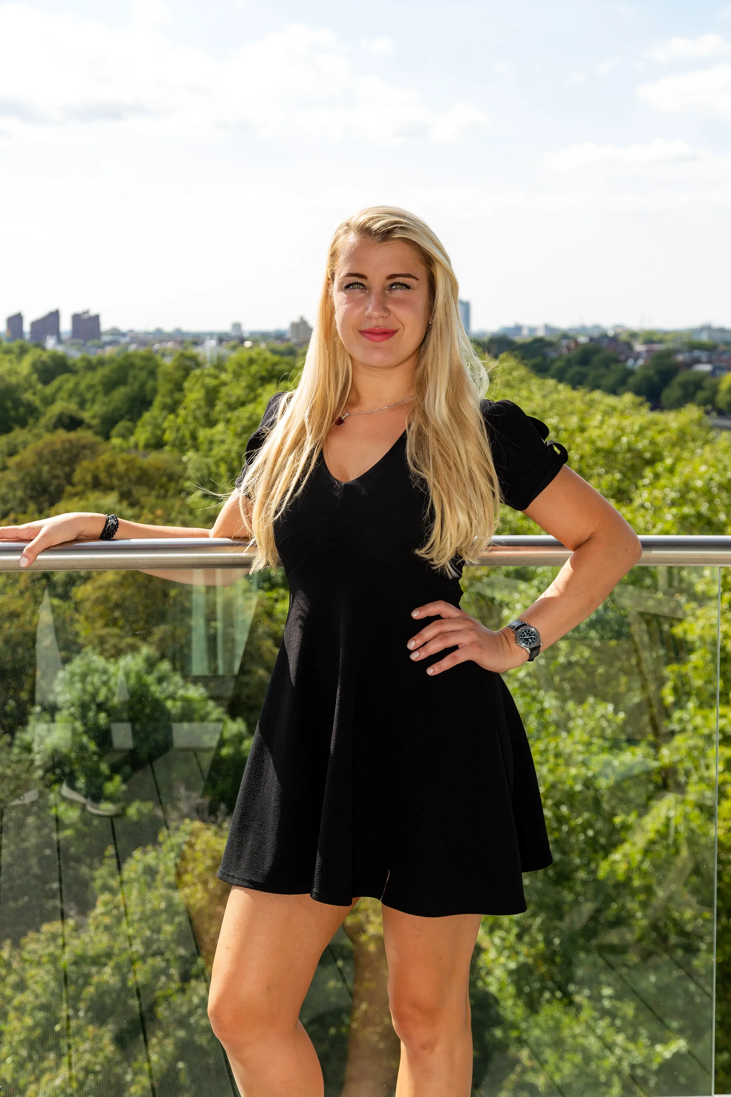 A young woman with long blonde hair wearing a black dress, standing outdoors on a balcony with a cityscape and green trees in the background.