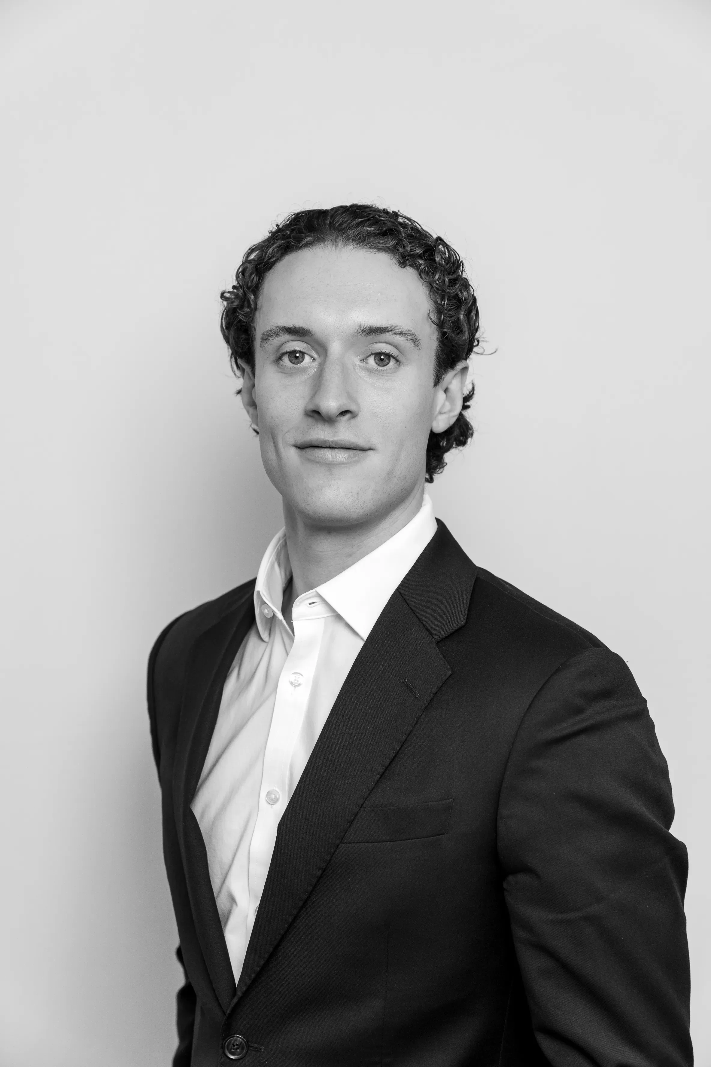 Black and white portrait of a young man with curly hair, dressed in a suit and white shirt, standing against a plain background.
