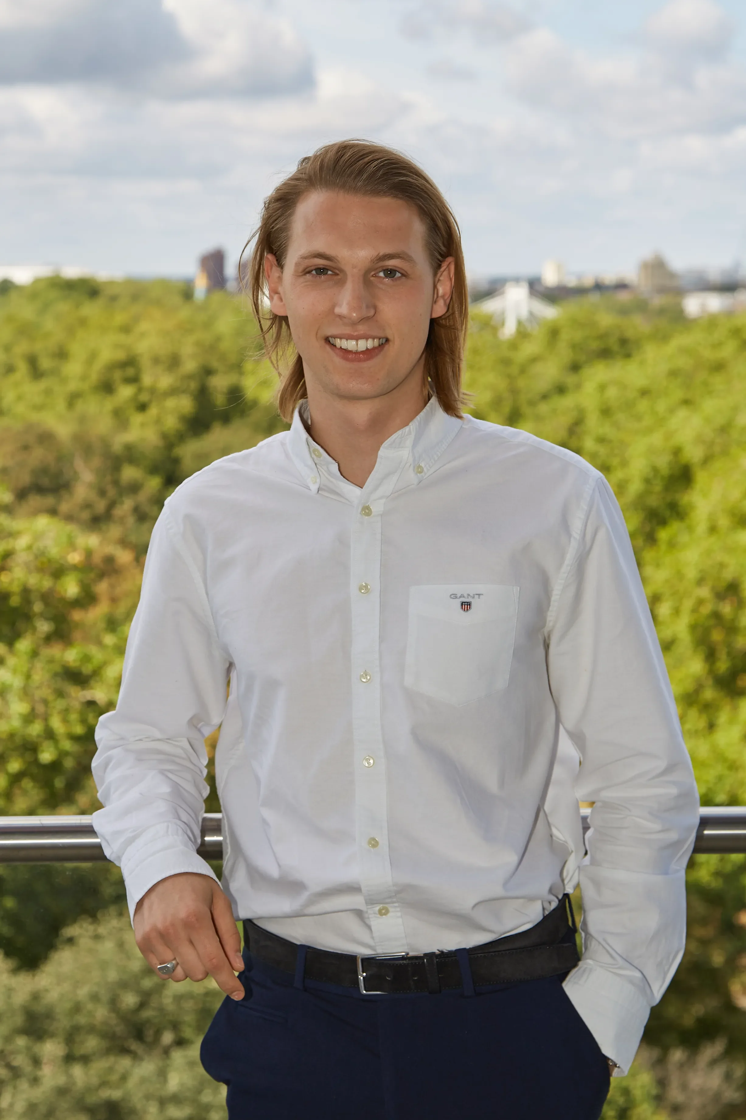 A young man with long hair, wearing a white dress shirt, standing outdoors with a green landscape and city buildings in the background, smiling at the camera.