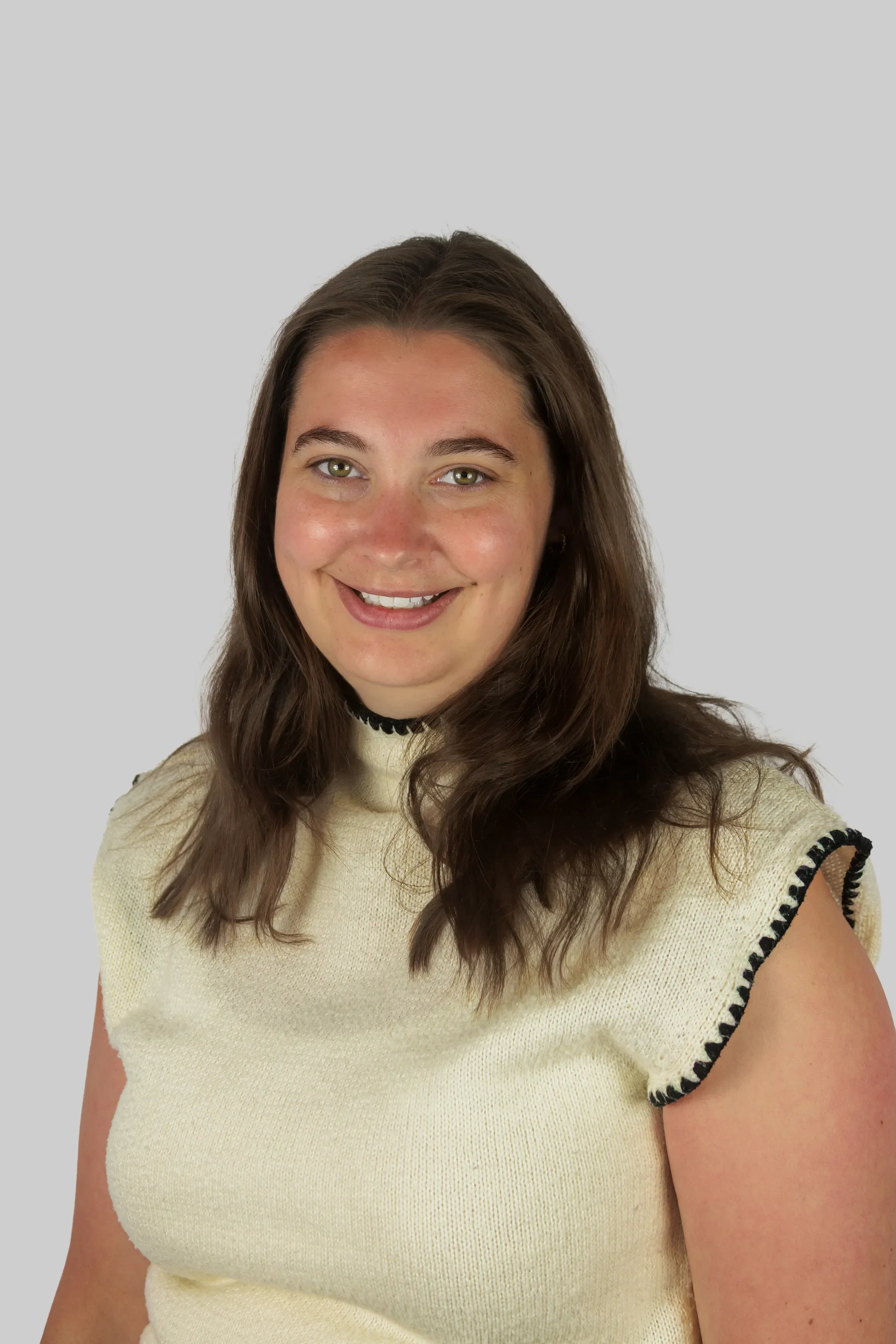 A young woman with brown hair and green eyes smiling, wearing a cream-colored knit top with black trim details.