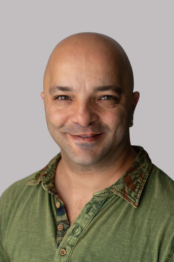 Bald man with light skin and blue eyes smiling, wearing a green shirt with a patterned collar, against a light gray background.