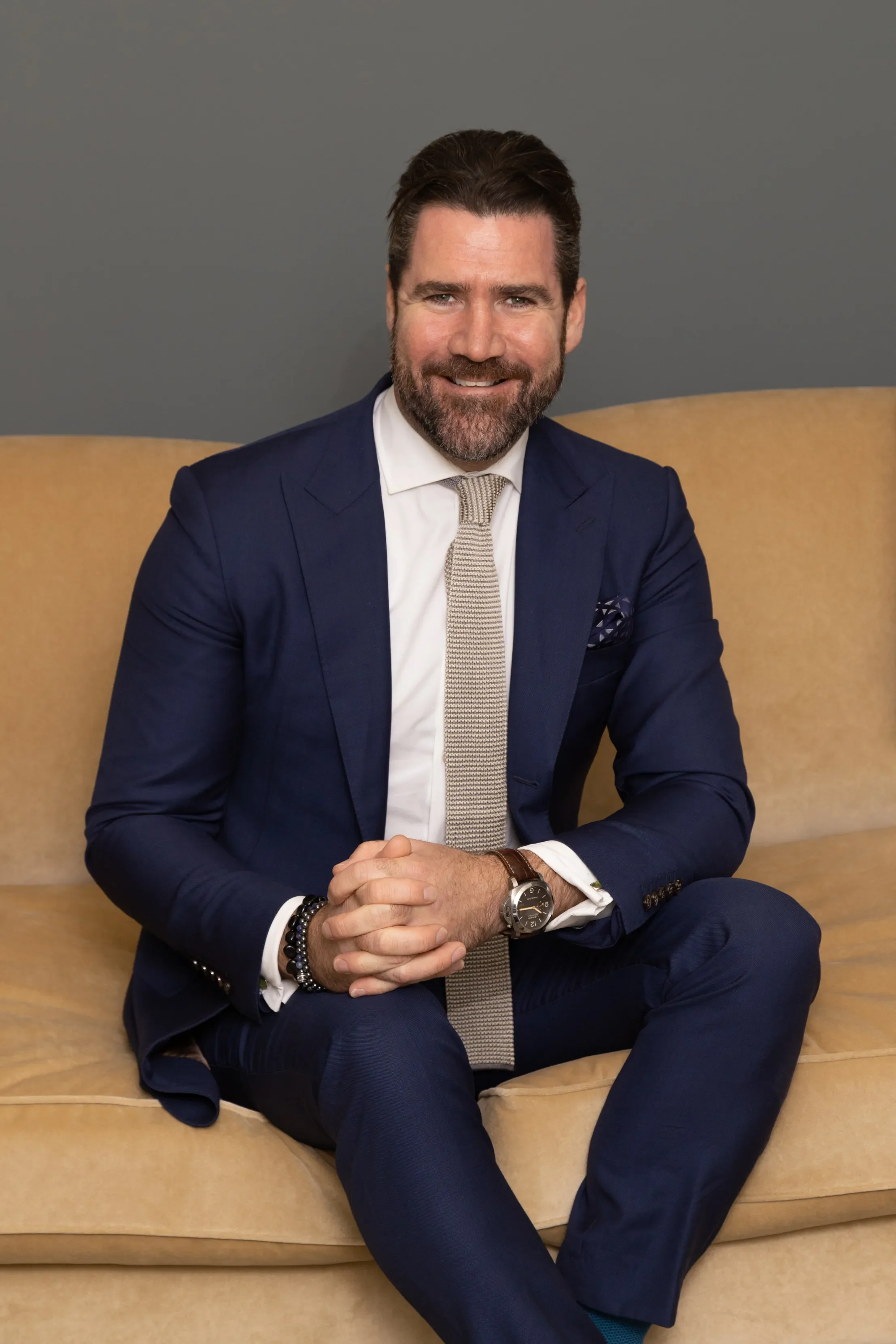 A man with dark hair and a beard, wearing a navy blue suit, white shirt, and light-colored tie, sitting on a beige sofa with a gray background, smiling at the camera.
