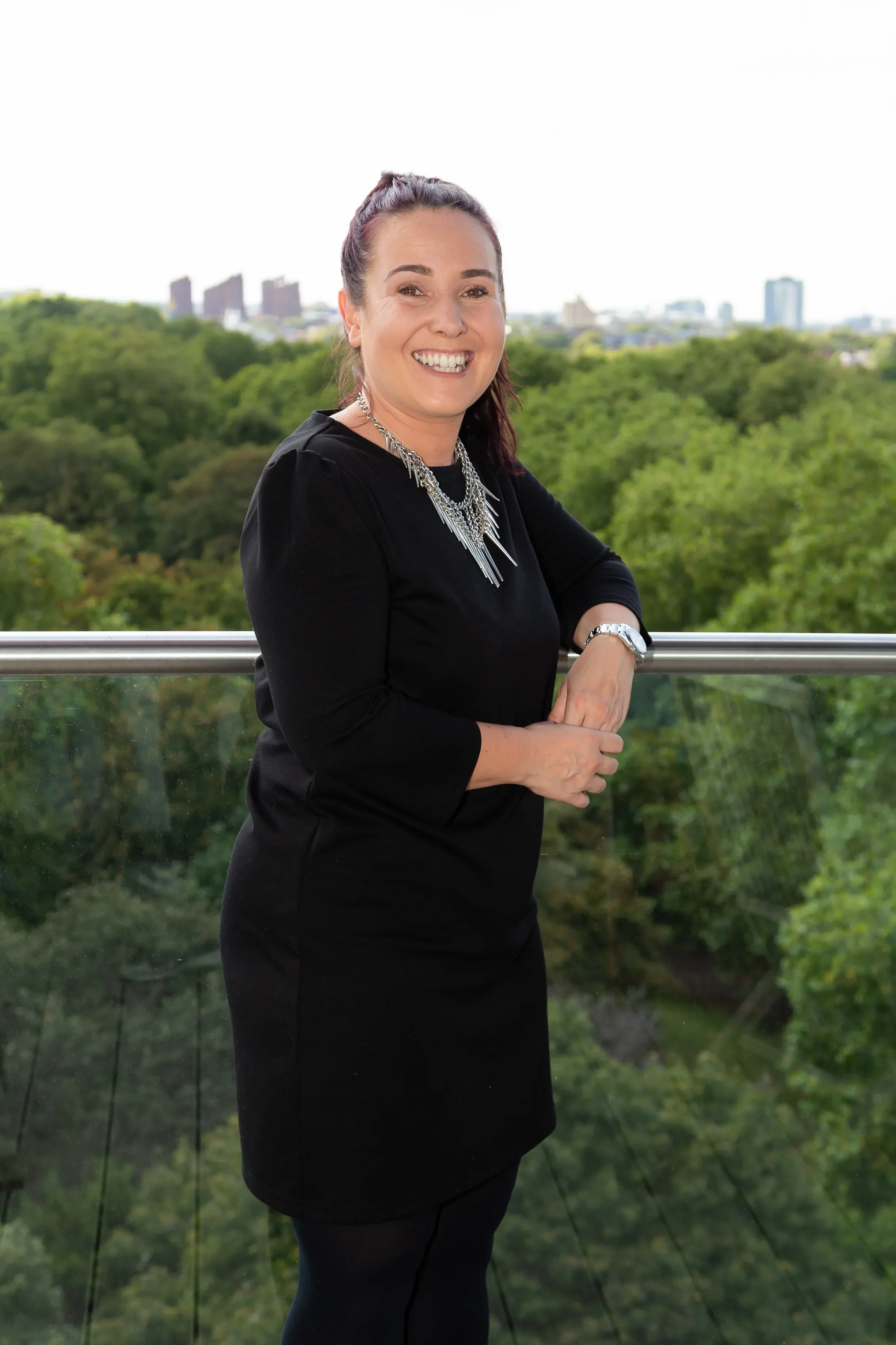Smiling woman in black dress with necklace and watch, standing outdoors on a balcony with green trees and city skyline in the background.