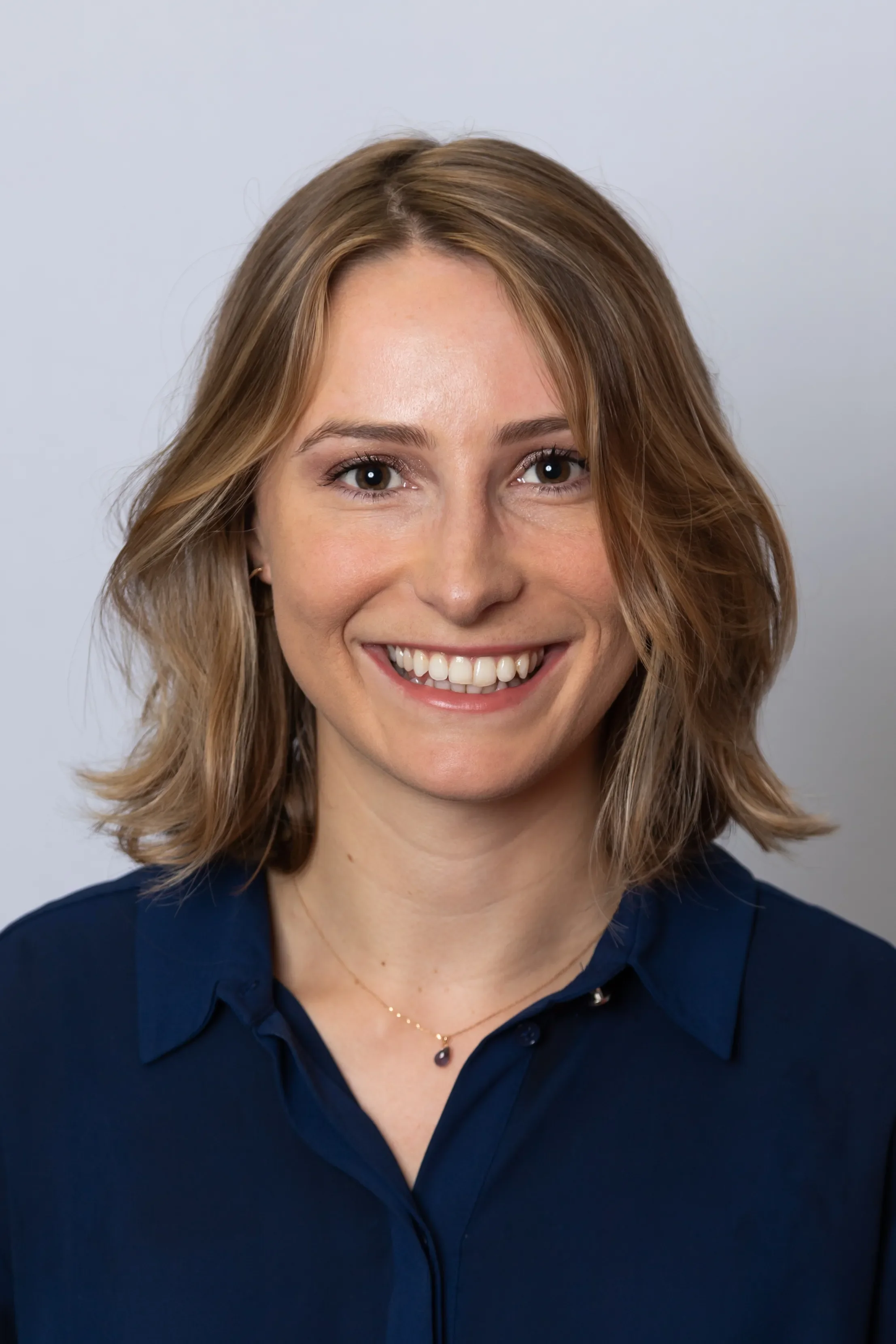 Close-up of a woman with shoulder-length light brown hair smiling, wearing a dark blue collared shirt and a delicate necklace, against a plain light gray background.