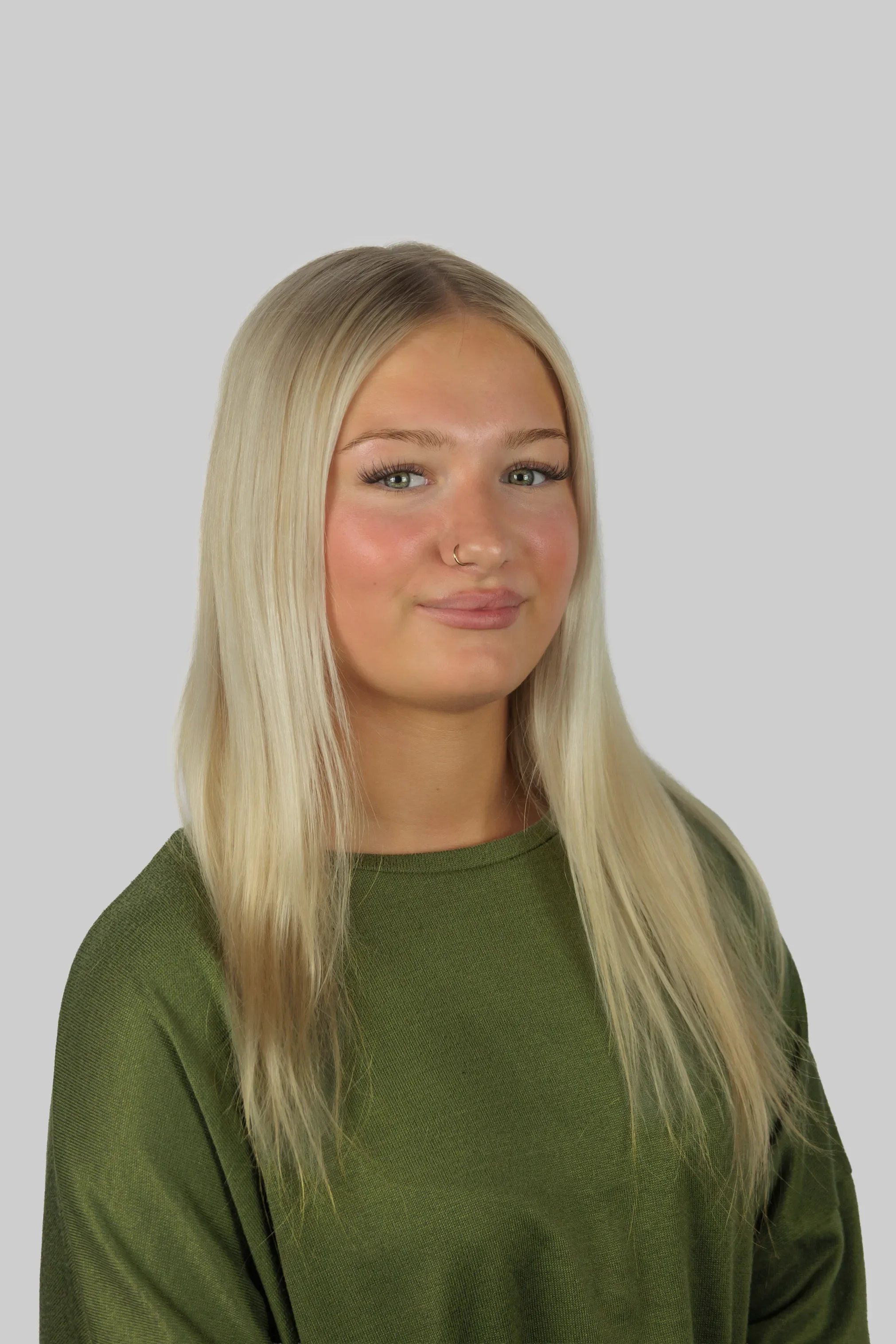 A young woman with long blonde hair, light skin, and a nose ring, wearing a green sweater, smiling softly against a plain light gray background.