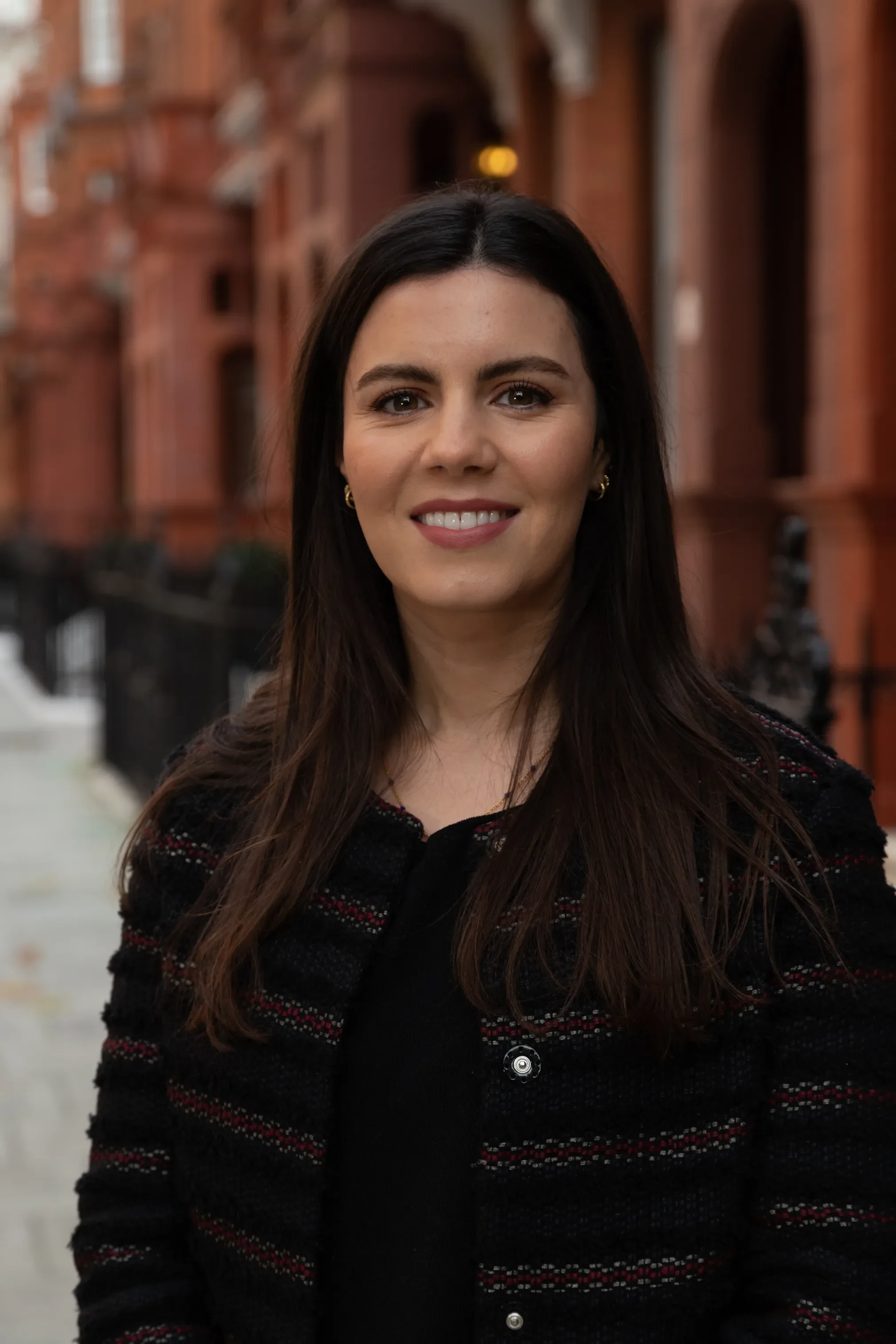 A woman with long dark hair, wearing a black and red patterned sweater, smiling outdoors in front of a red brick building.