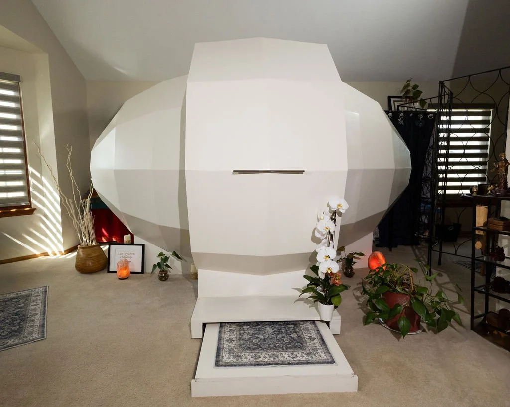 A large white geometric structure made of folded paper or material, resembling a spaceship or capsule, is placed indoors on a small platform with a rug. It is surrounded by houseplants and decor, with sunlight streaming through the window blinds.