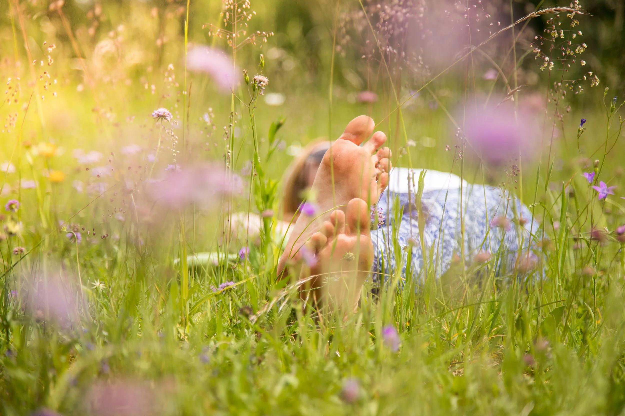 Person lying on their back in a grassy field with wildflowers, holding their feet with both hands.