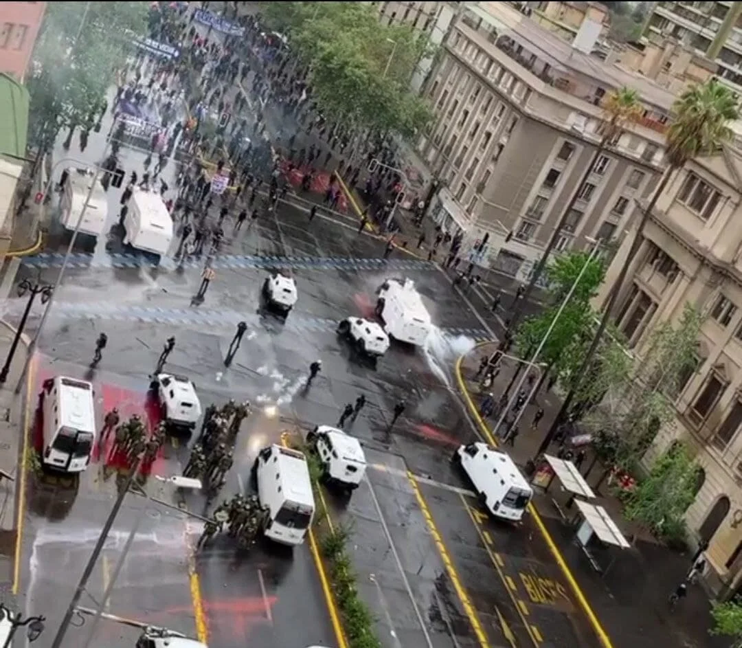 Momentos cuando un sujeto encapuchado a la derecha de la imagen, debajo del semáforo, lanza un explosivo que impacta en la abogada Denisse Cortés Saavedra, quien estaba hablando con Carabineros.